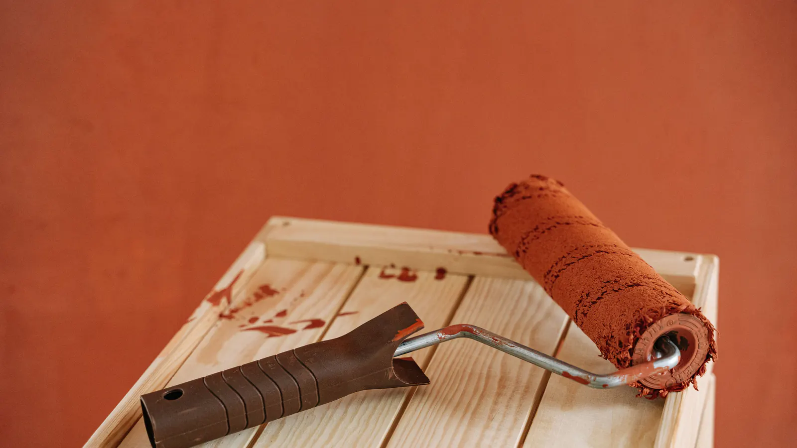 A paint roller loaded with reddish-brown stain resting on light wooden boards, brown streaks on the wood, against a terracotta wall