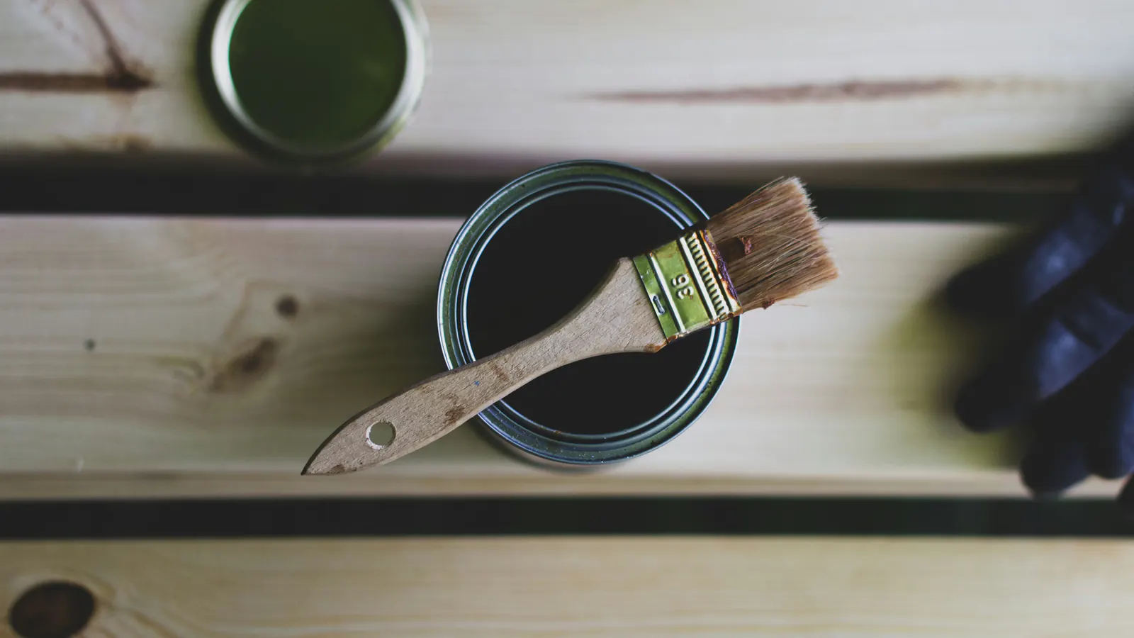 A natural-bristle brush laid across an open can of dark wood finish on unfinished pine boards, with a gloved hand at the edge