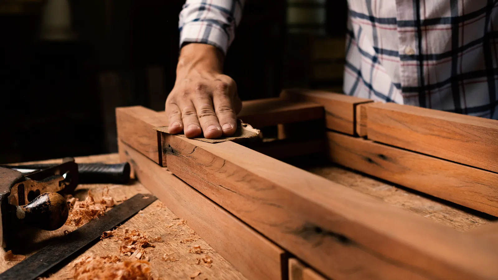 A hand pressing sandpaper along a reddish wood frame piece on a workbench scattered with shavings, a hand plane beside it