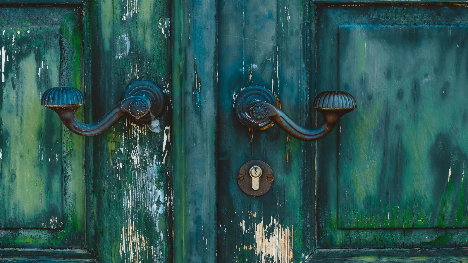 Close view of a paneled wood double door with cracked, peeling dark green paint and bare wood showing through, with curved iron handles