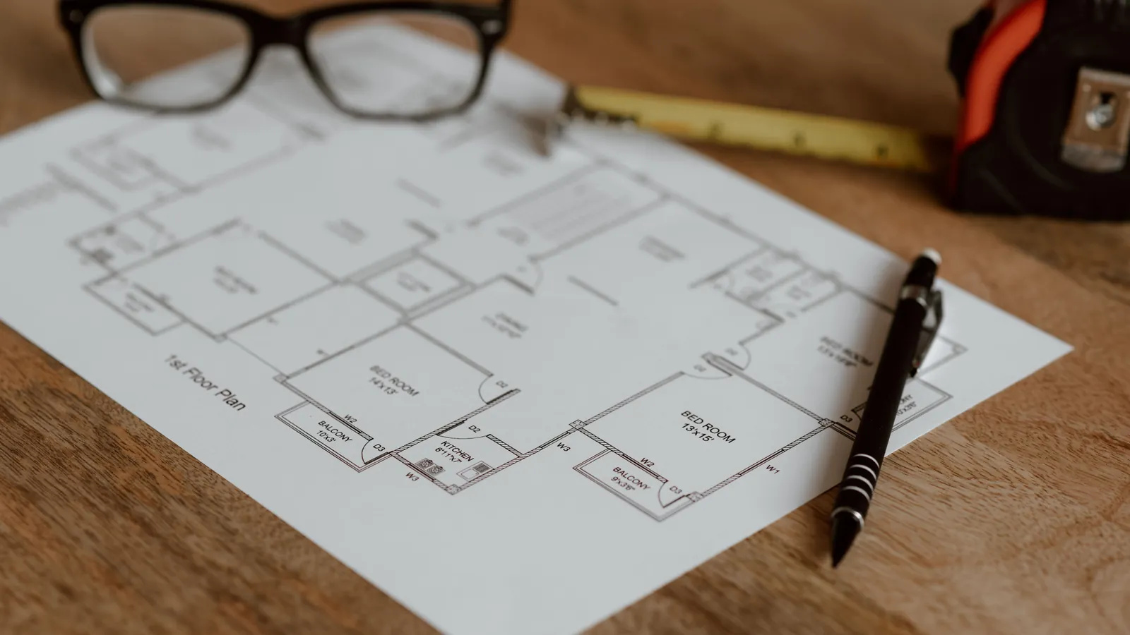 A printed house floor plan on a wooden table with a pen, reading glasses and a tape measure
