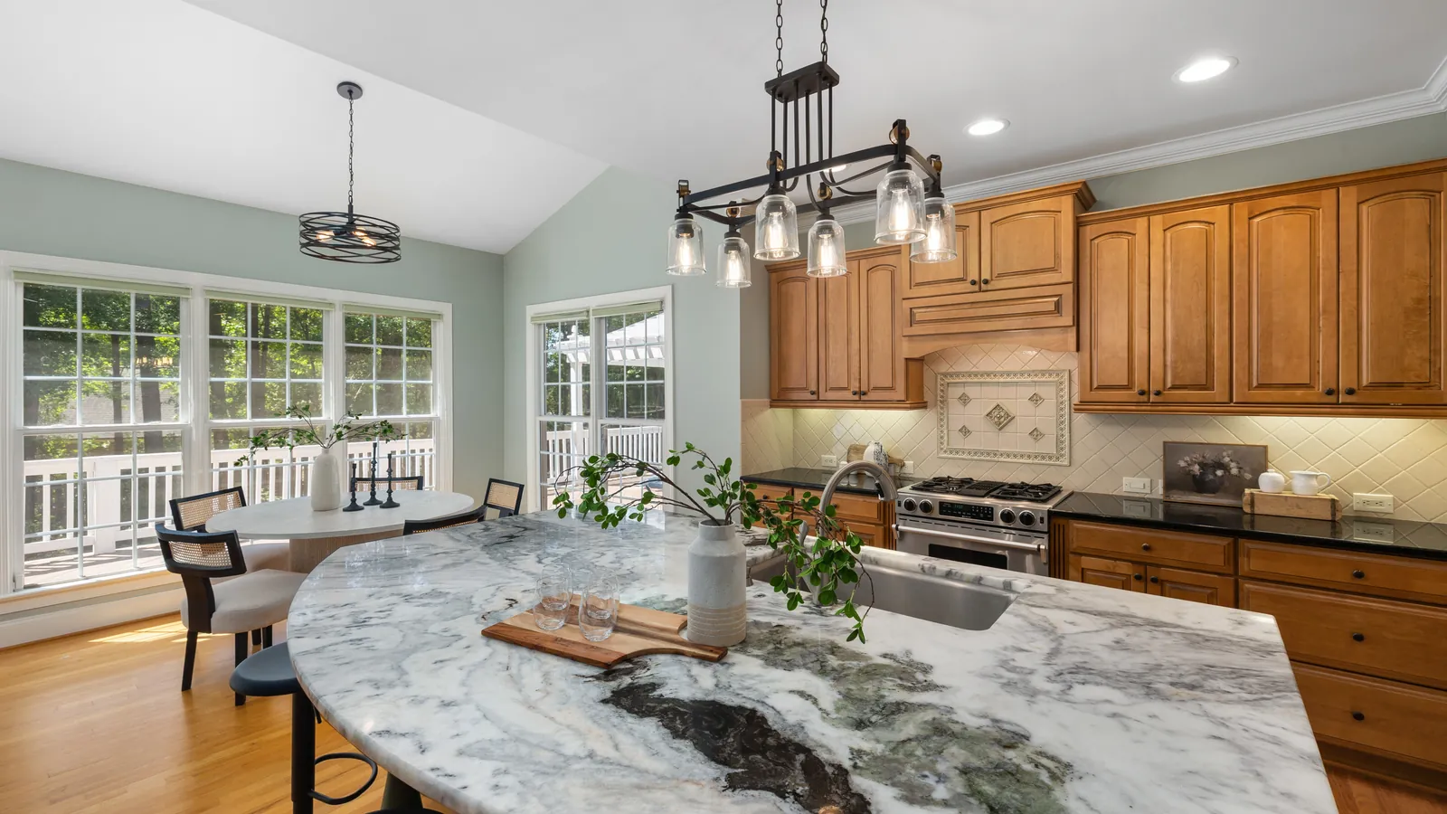 A kitchen with a curved marble island and sink, wood cabinets along the wall and a breakfast table by tall windows