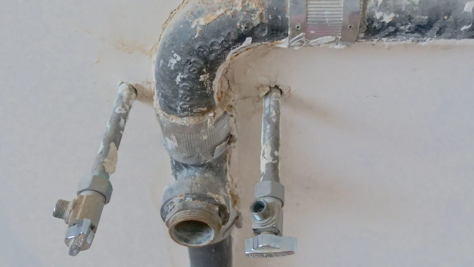 Old gray water pipes and shutoff valves coming out of a white wall where a fixture was removed