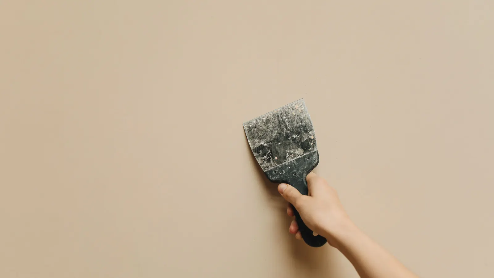 A hand holding a worn metal scraper flat against a smooth beige painted wall, the blade marked with old dried filler