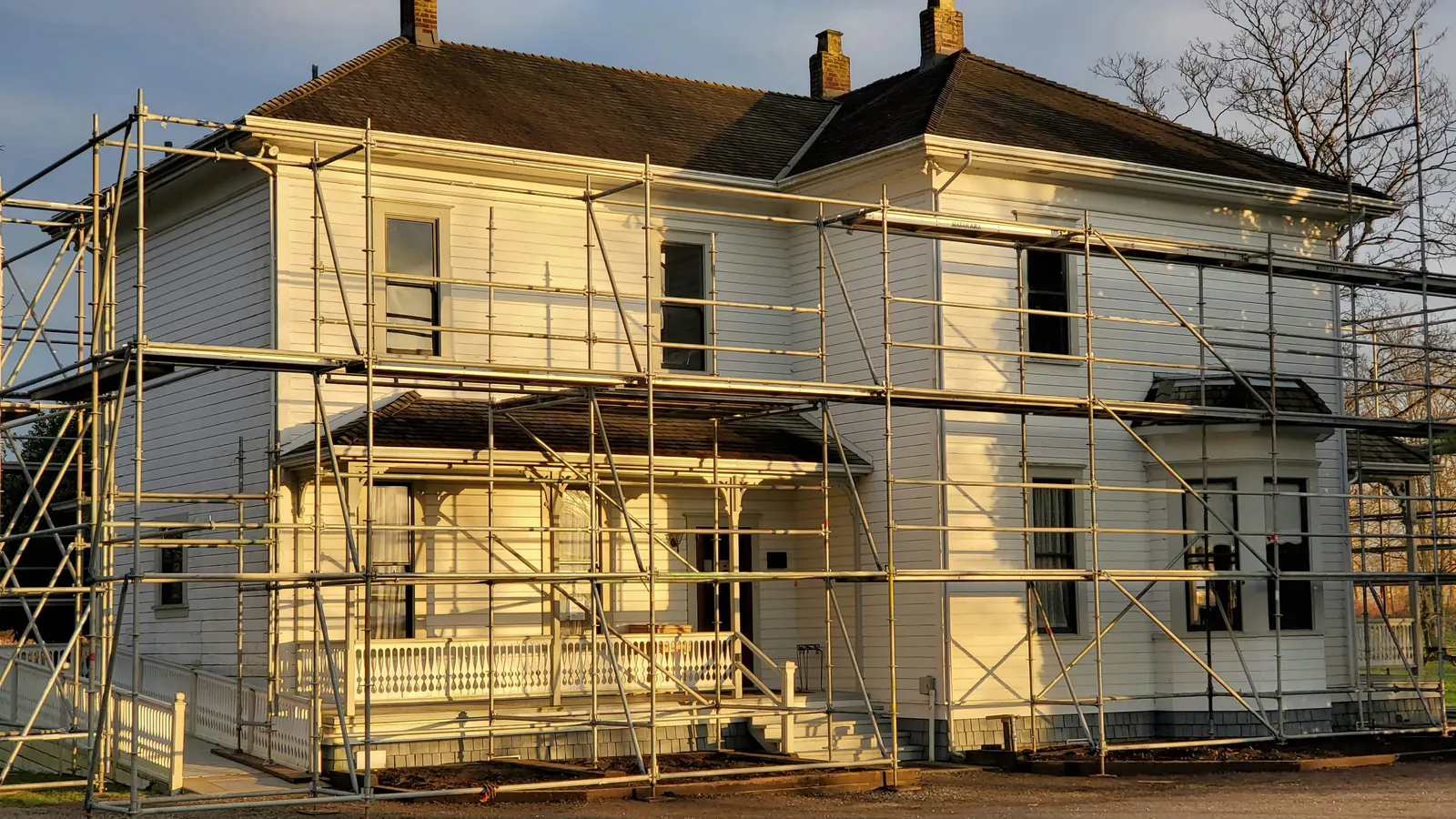 A two-story white clapboard house with tall chimneys and a front porch, surrounded on all sides by metal scaffolding in warm evening light