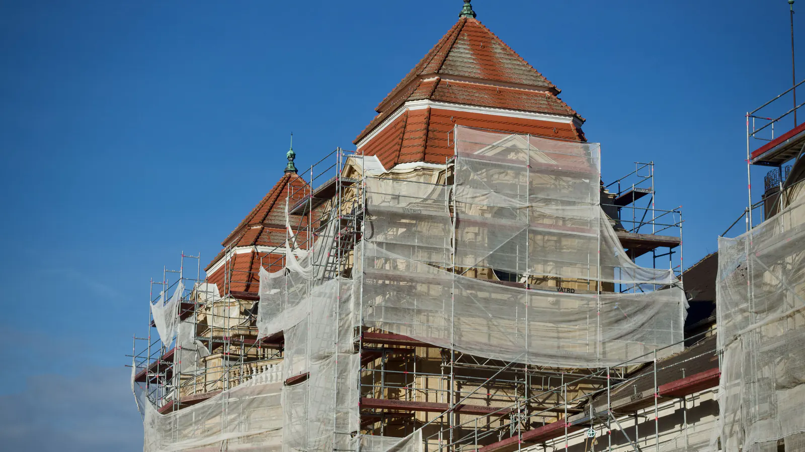 Scaffolding around a historic building with tiled towers, wrapped in translucent protective sheeting against a deep blue sky