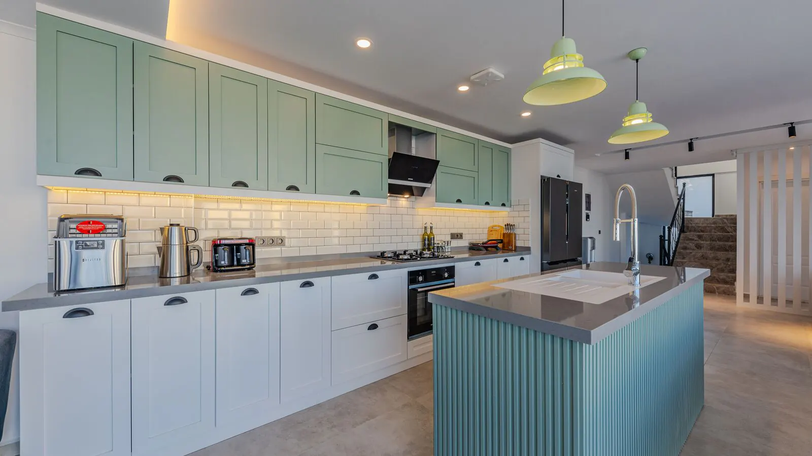 A long kitchen with sage green upper cabinets, white lower cabinets, a white subway tile backsplash and a pale blue island under two pendant lights