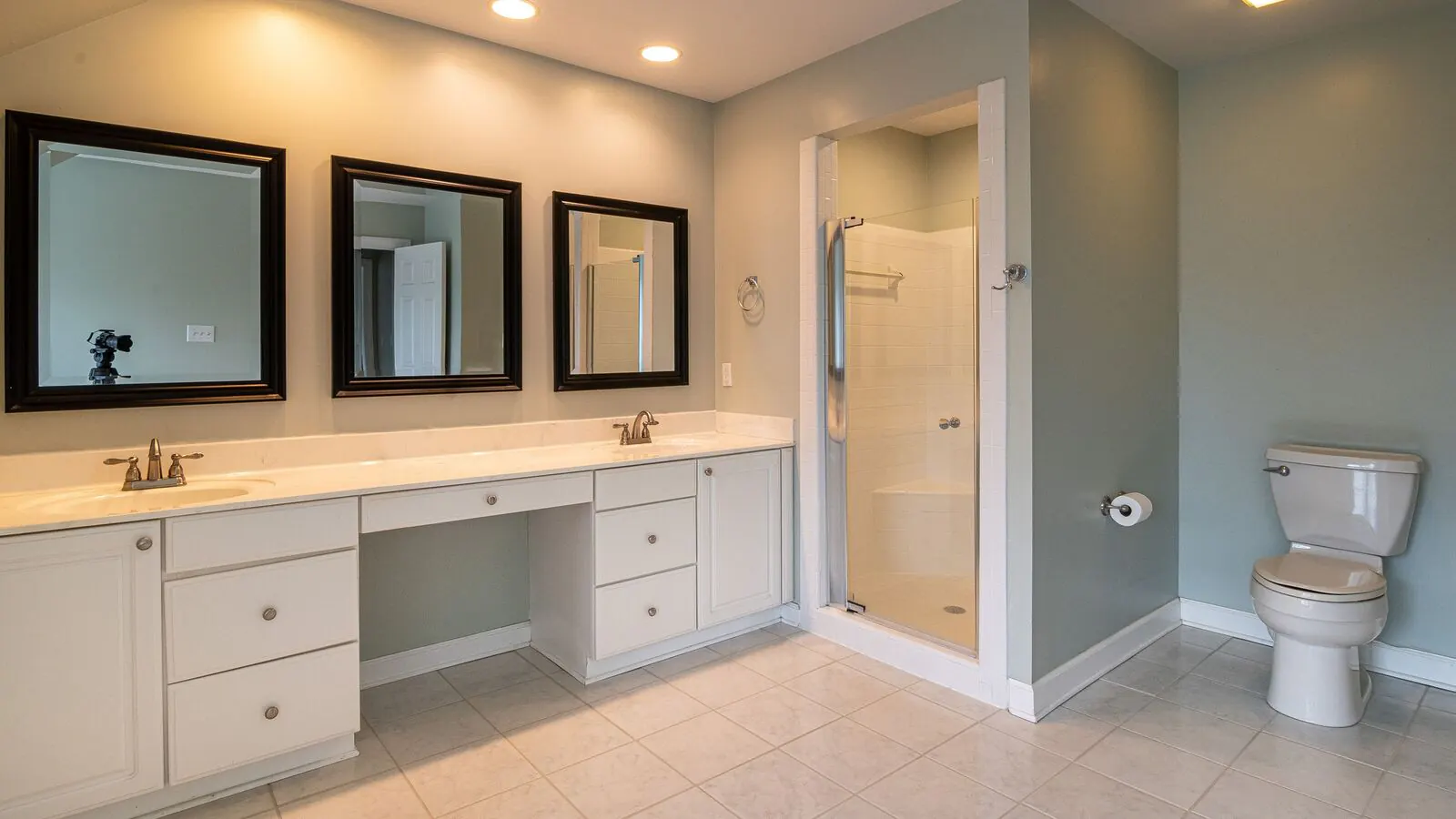 A bathroom with gray-green painted walls, a long white vanity with three mirrors above it and a shower stall beside a toilet