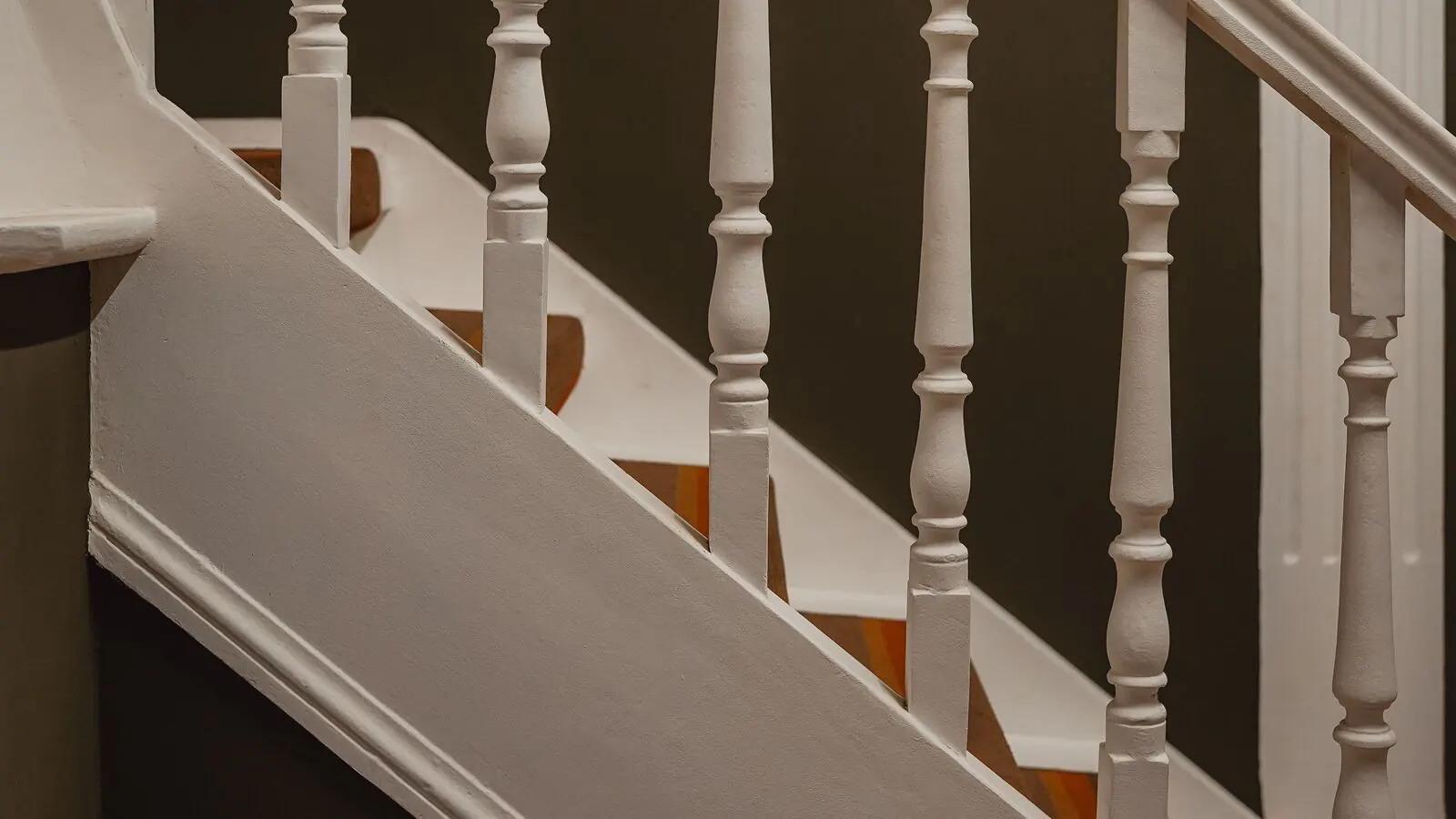 White painted turned balusters, handrail and stringer on a stair against a dark green wall