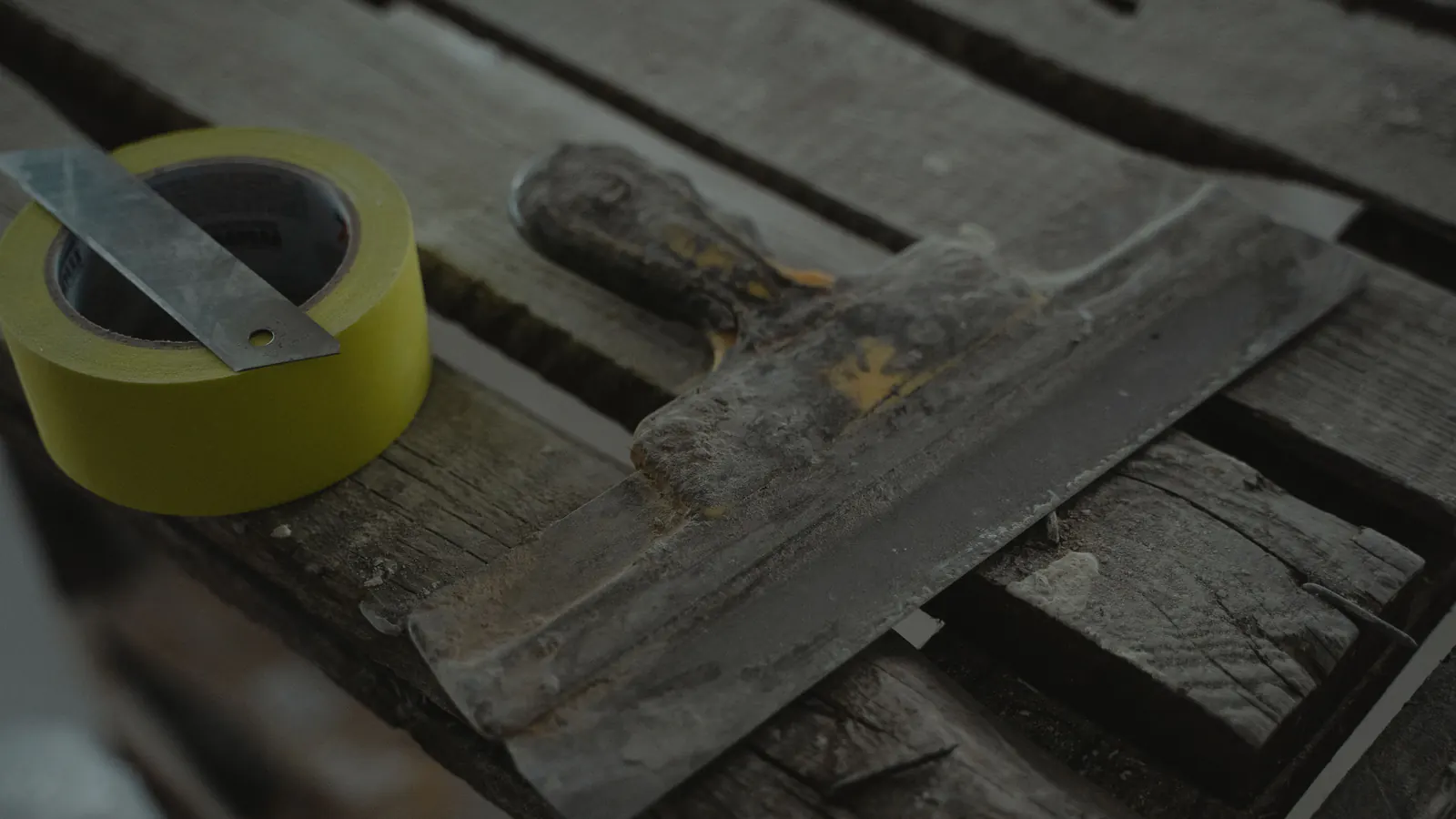 A worn putty knife crusted with filler beside a roll of yellow tape and a metal ruler on weathered gray boards