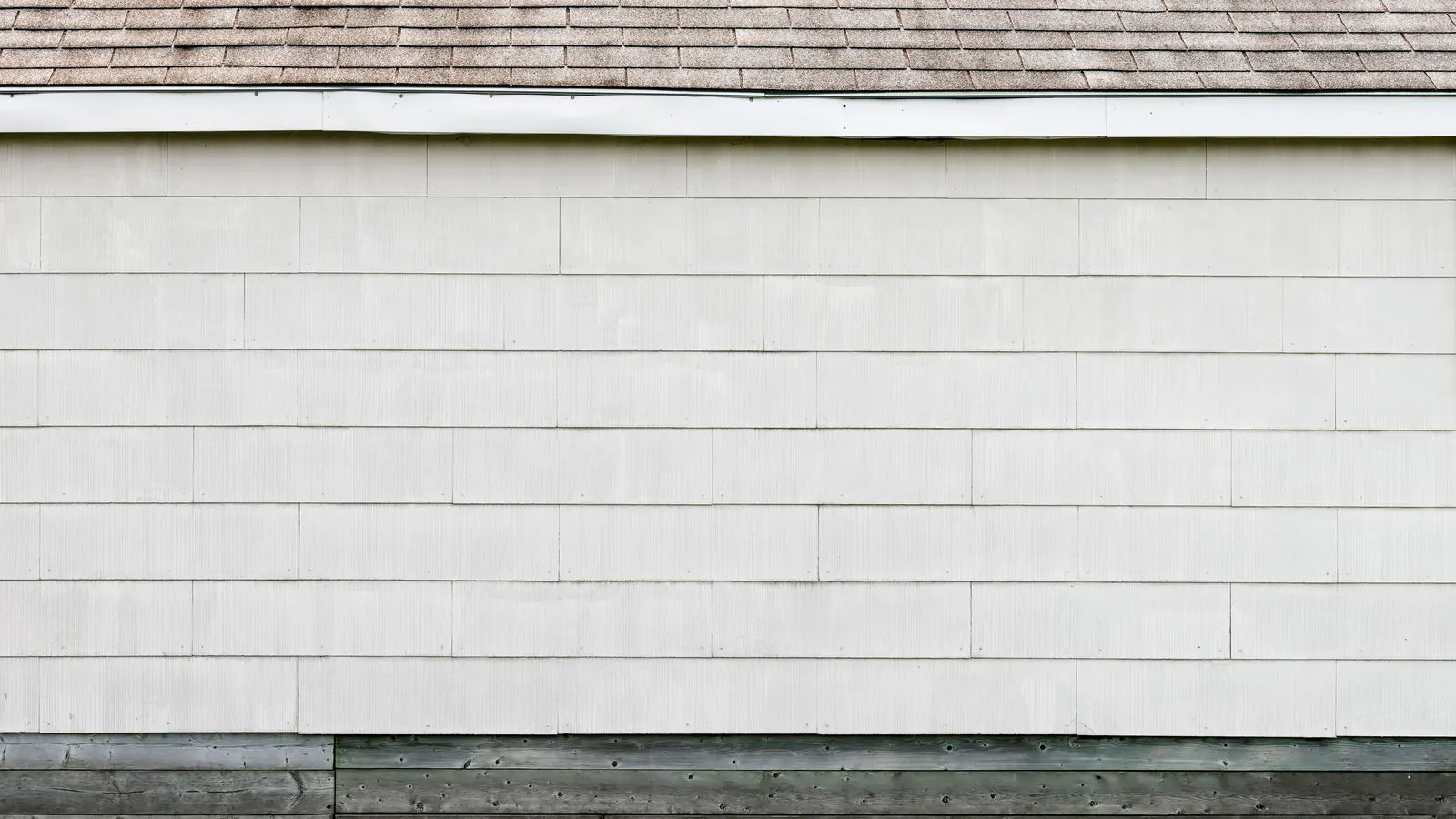A wall of white painted shingle siding meeting a white trim board under an asphalt shingle roof edge, gray boards along the base