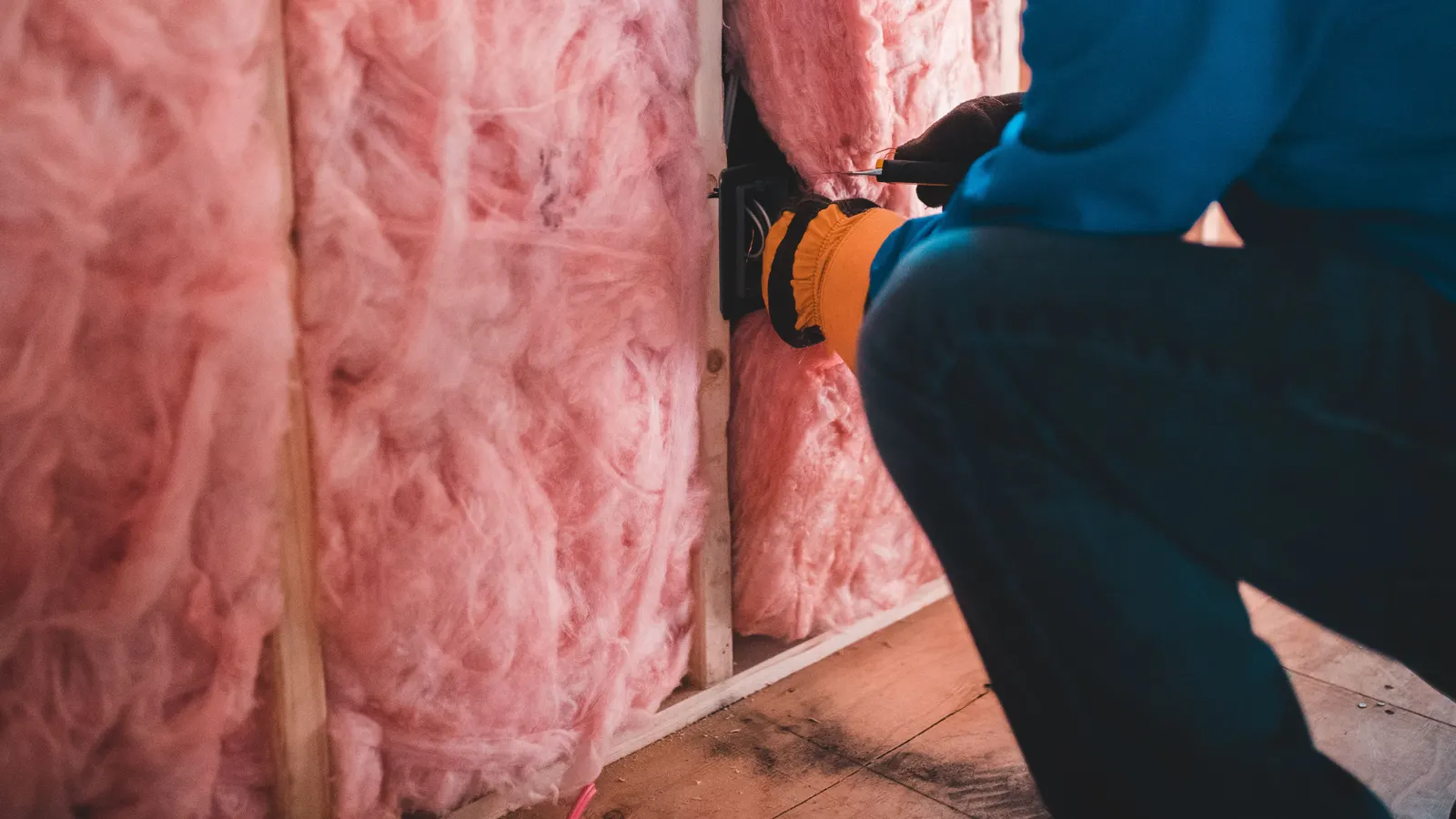 A gloved worker, face out of frame, fitting pink insulation batts between wall studs
