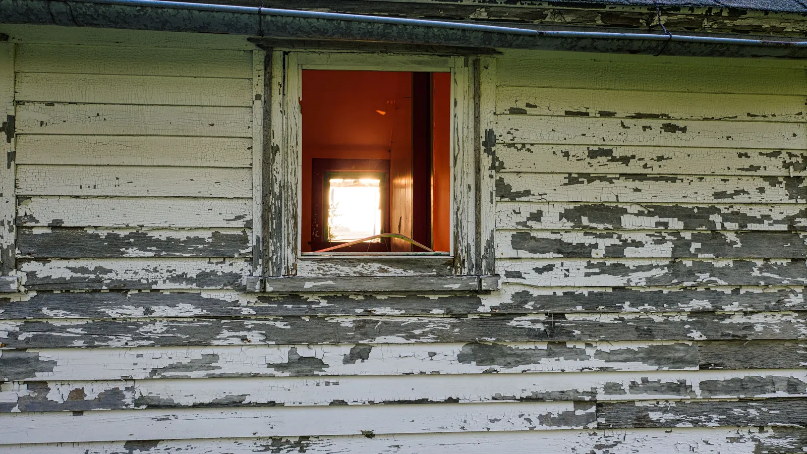 Worn white paint peeling from wood lap siding around a small window
