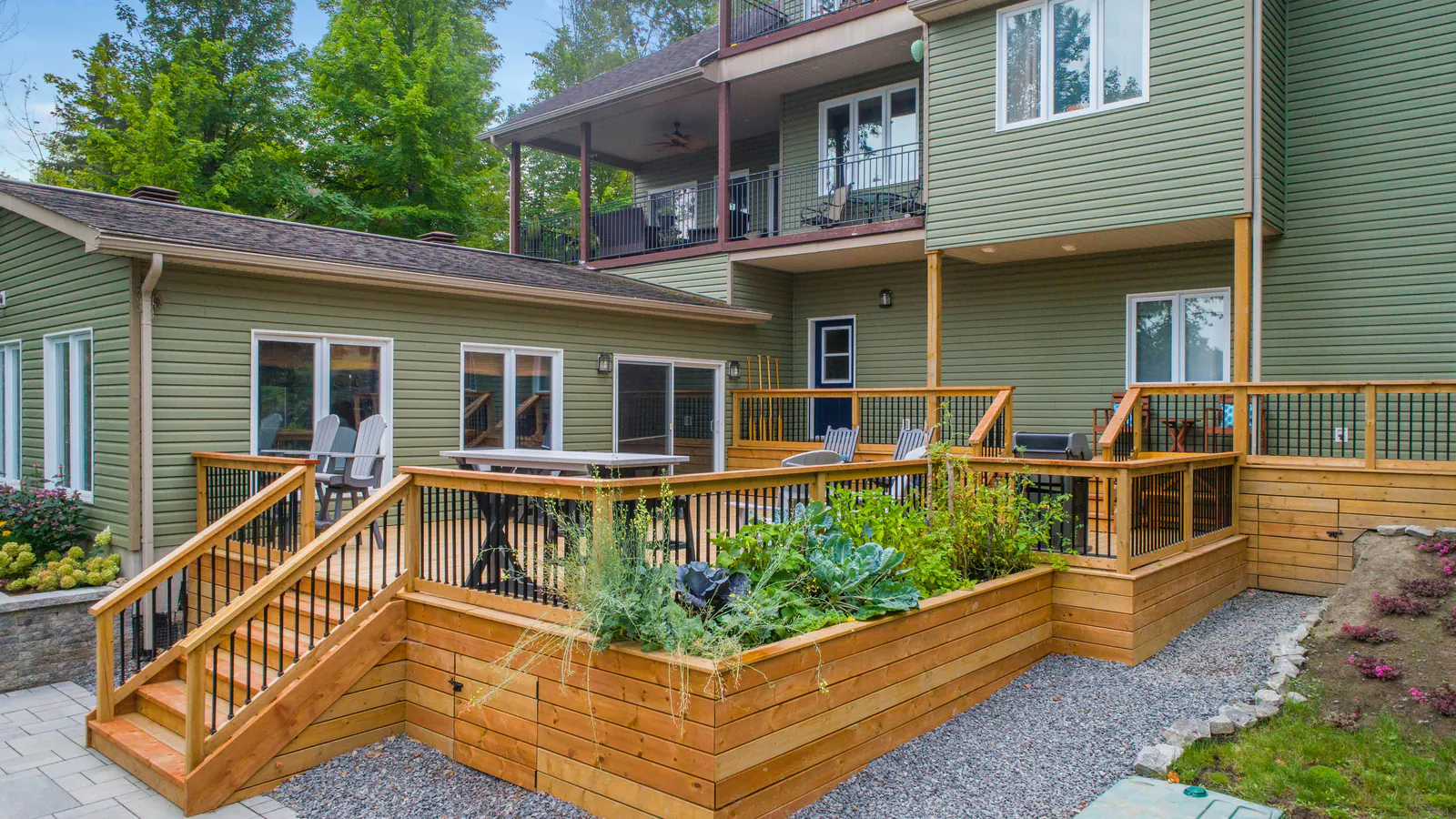 A newly built natural wood deck with stairs, railings and raised planters behind a large green lap-sided house with trees around it.