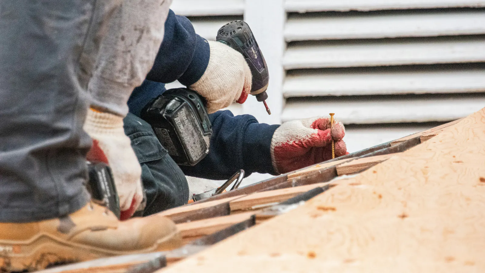 Gloved hands drive a fastener with a cordless drill into fresh plywood roof decking along the eave, next to a white louvered vent.
