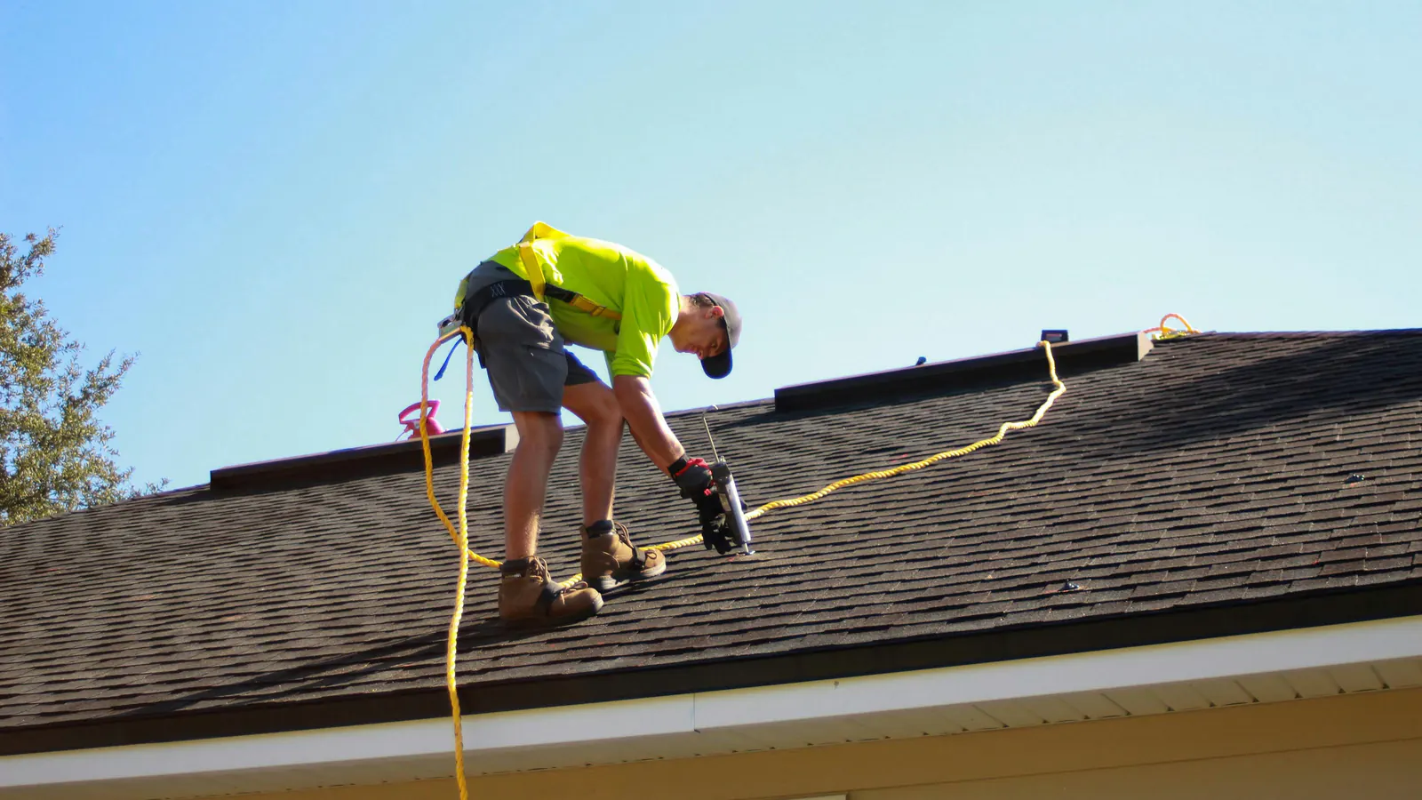 A roofer in a safety harness and bright shirt bends over a dark asphalt shingle roof working with a caulk gun under a clear blue sky.