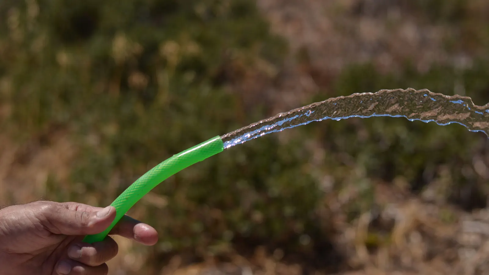 A hand holding a bright green garden hose with a strong stream of water arcing out against a blurred dry hillside