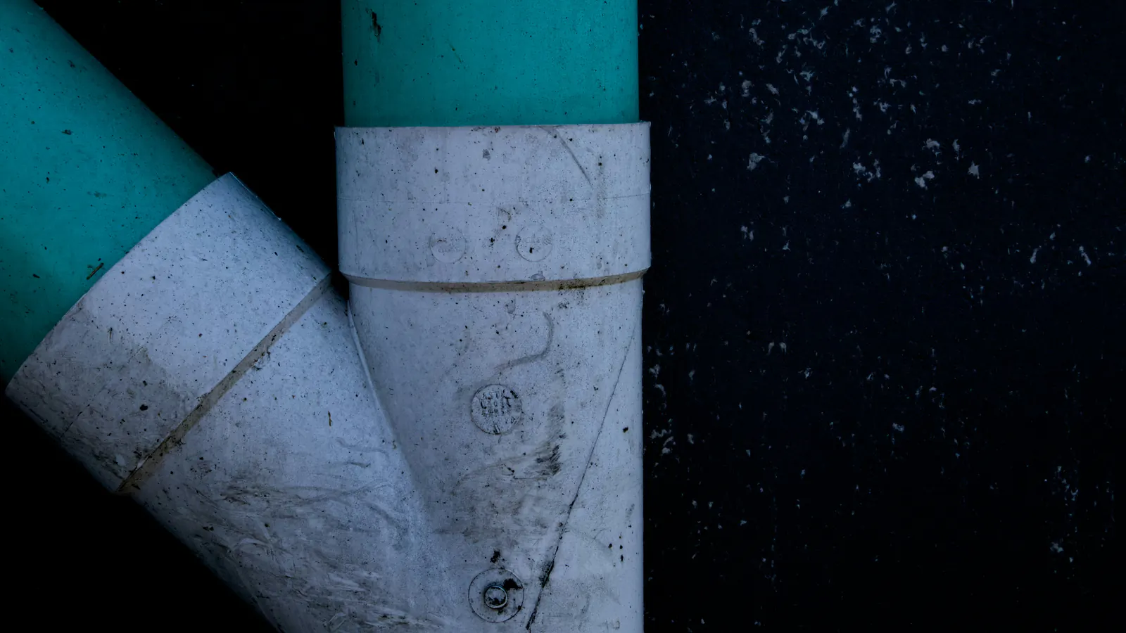A white PVC wye fitting joining two teal plastic pipes against a dark background, with scuffs and markings on the fittings