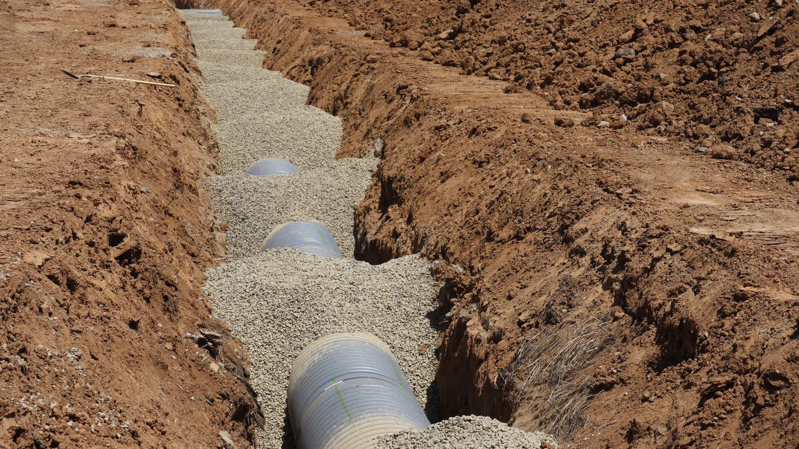A corrugated drainage pipe laid in a bed of pale gravel at the bottom of a freshly dug trench of red-brown soil