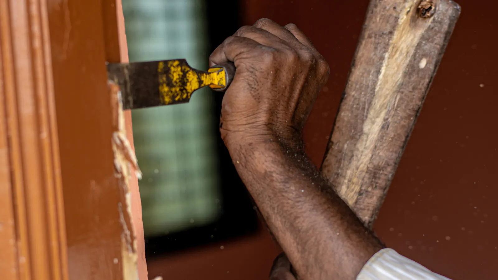A hand working a chisel against a painted wooden door frame, another hand gripping a wooden mallet handle