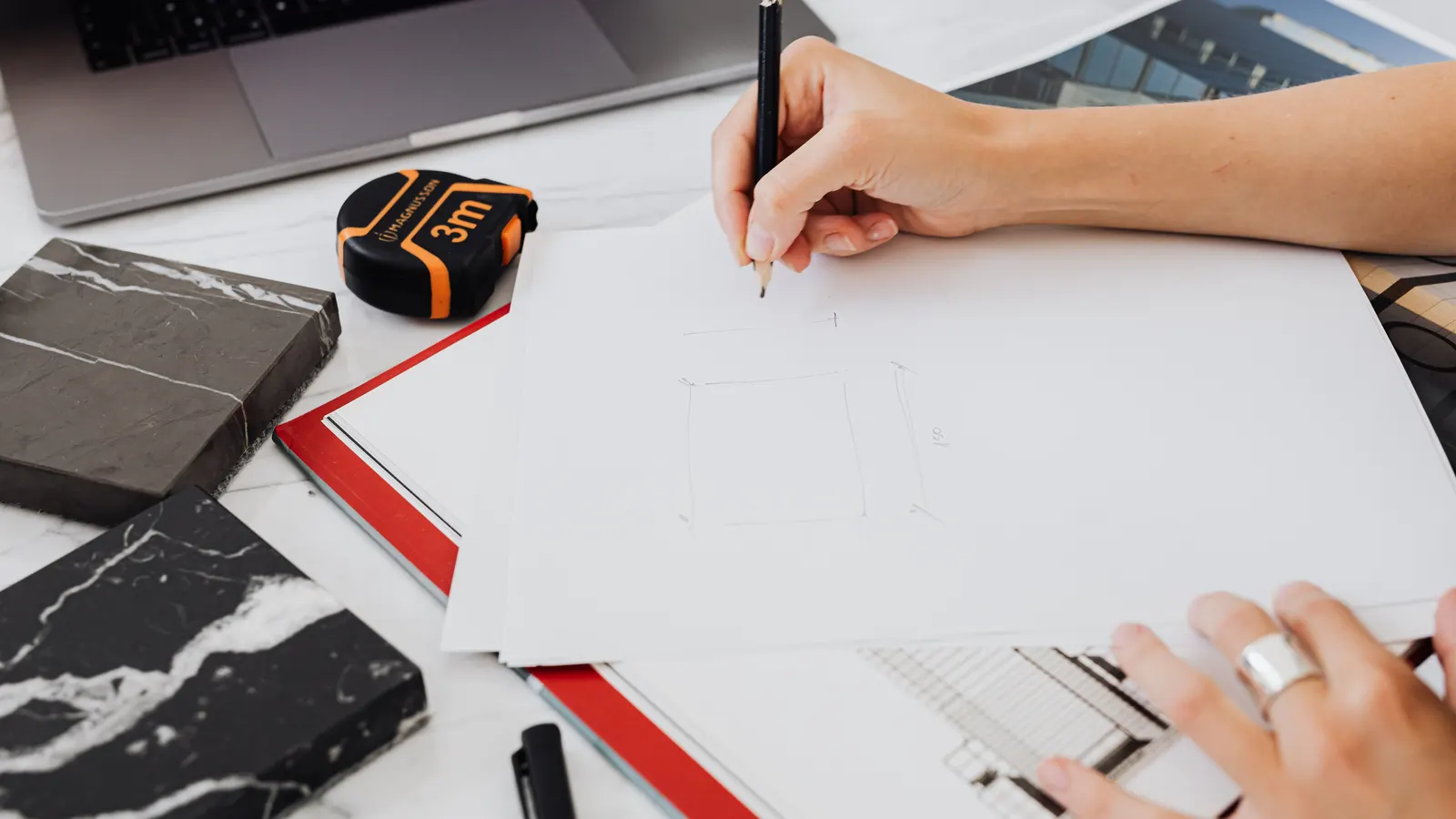 Hands sketching on drafting paper beside marble samples, a tape measure and a laptop