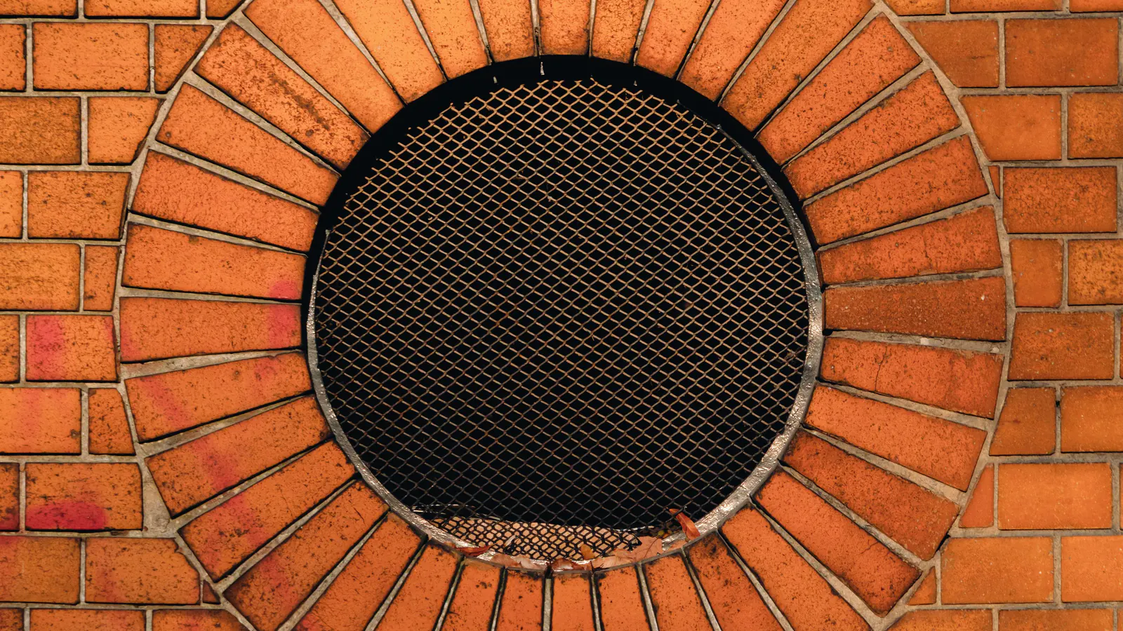 A round vent opening in a brick wall, covered with dark expanded metal mesh and ringed with bricks