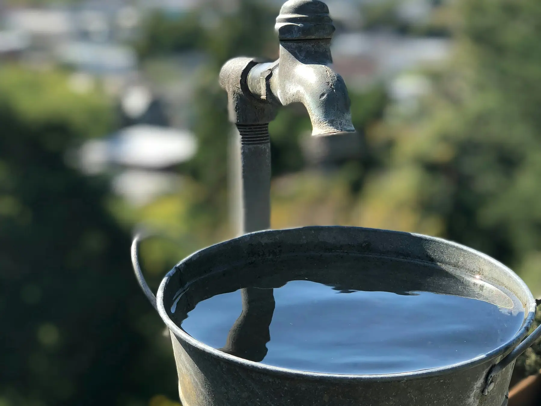 A galvanized bucket filled to the brim under an old outdoor faucet, hills blurred behind