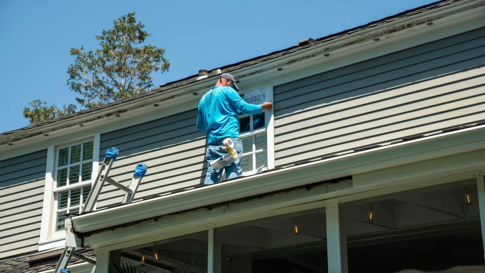 A worker in a blue shirt standing on a low porch roof where it meets gray lap siding, working at an upstairs window, seen from behind