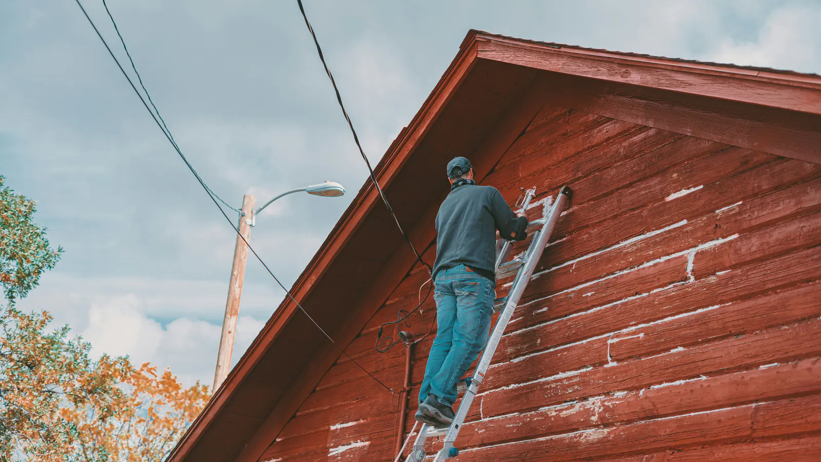 A painter seen from behind on an aluminium ladder coating weathered red wood siding on a gable wall under an overcast sky