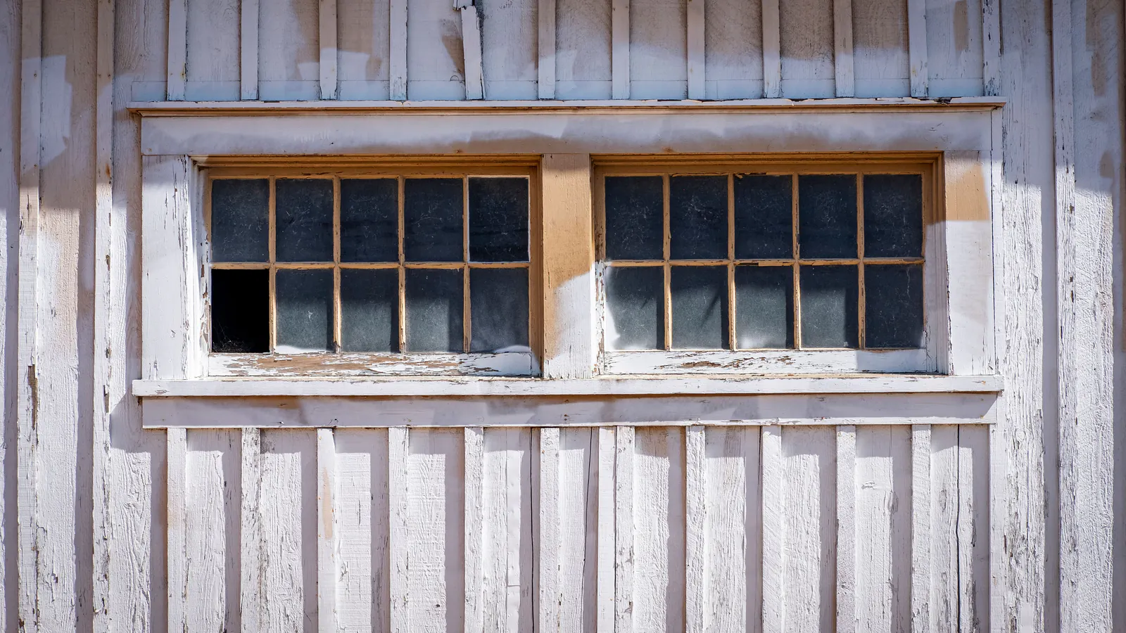 Two divided-light windows in a white board wall, the sill and frames cracked and peeling to reveal gray weathered wood