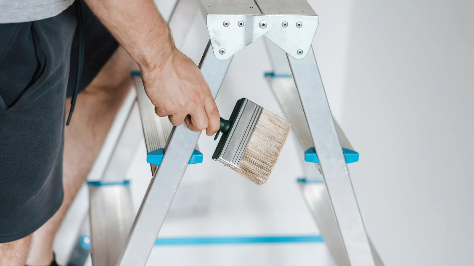 A hand gripping a wide paint brush beside an aluminium stepladder in a bright white room