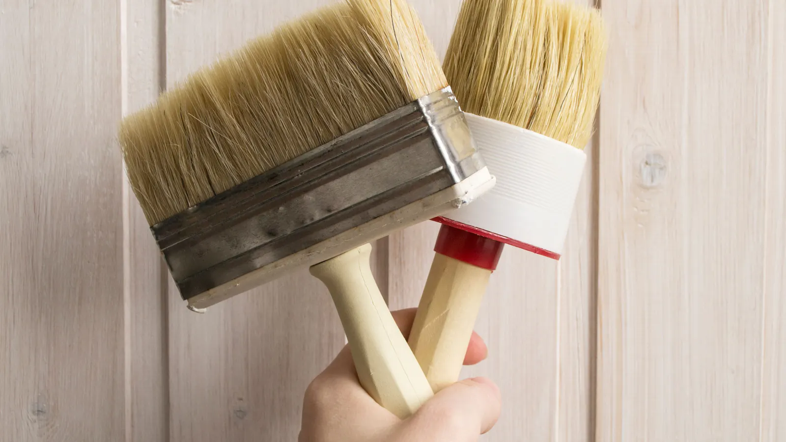 A hand holding two natural-bristle paint brushes, one flat and one round, in front of pale white-painted wood boards