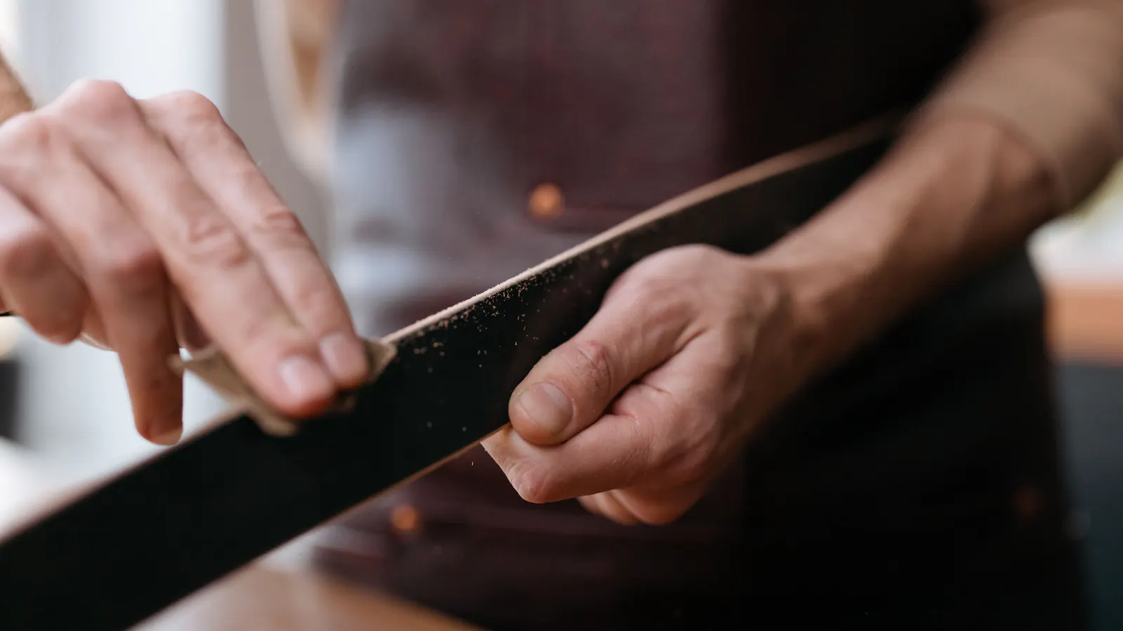 Close view of two hands working a folded piece of sandpaper along a dark wooden strip, fine dust lifting from it