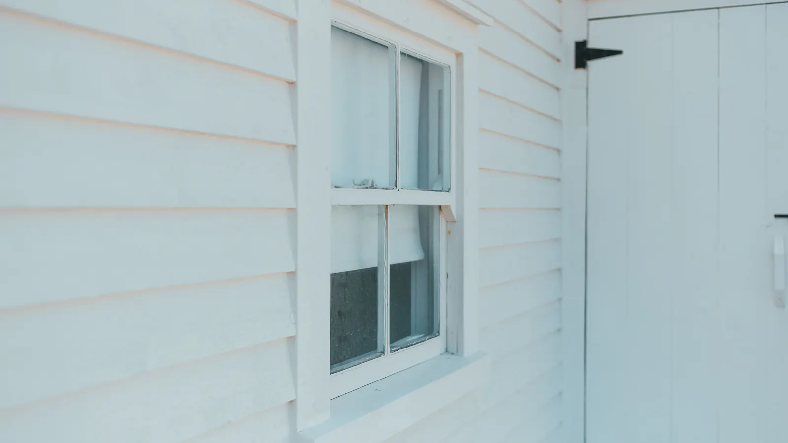 A freshly painted white double-hung wood window set in white lap siding beside a white board door with a black strap hinge