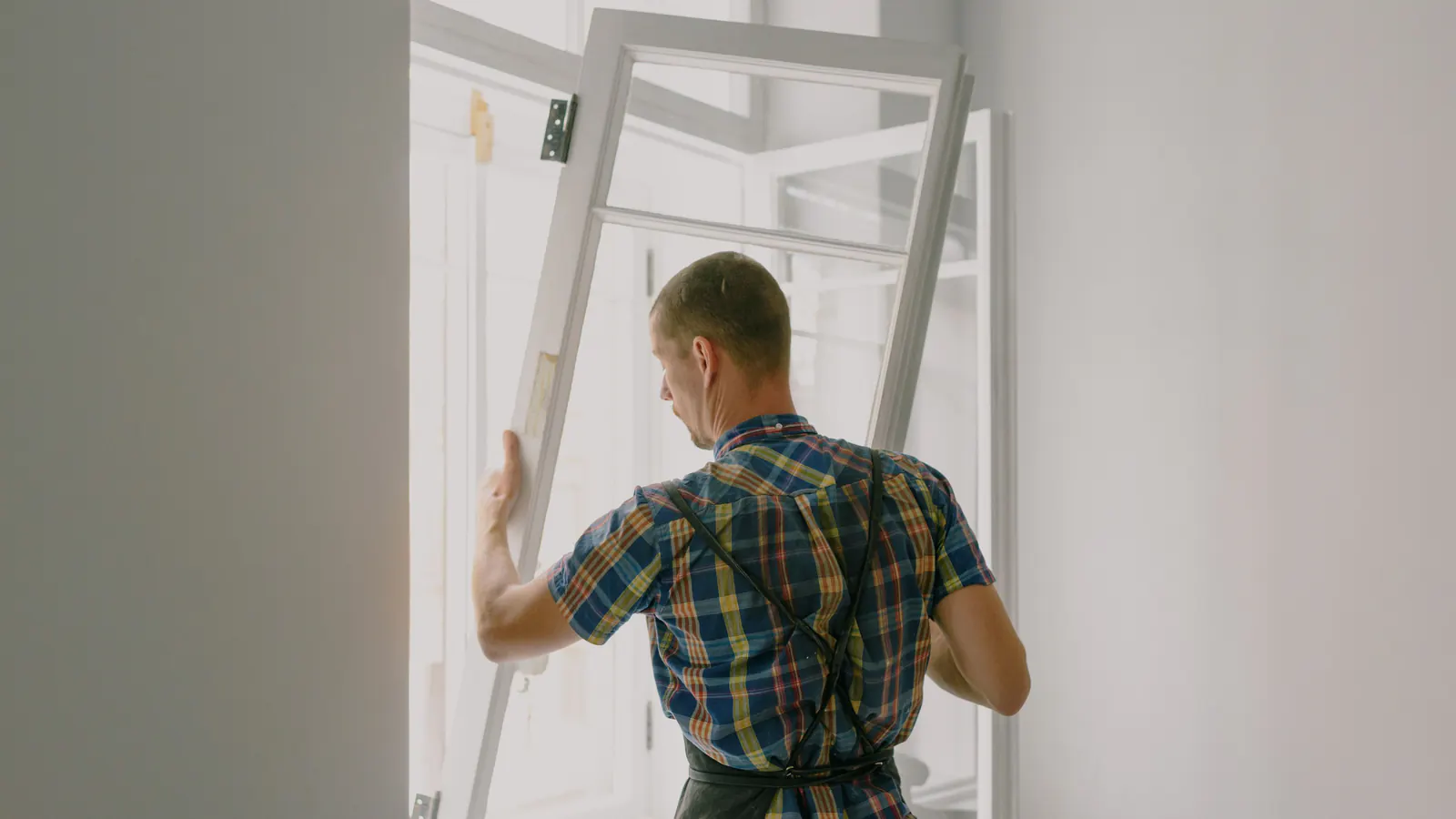 A man seen from behind in a plaid shirt and work apron lifting a white wood window sash into its frame in a bright room
