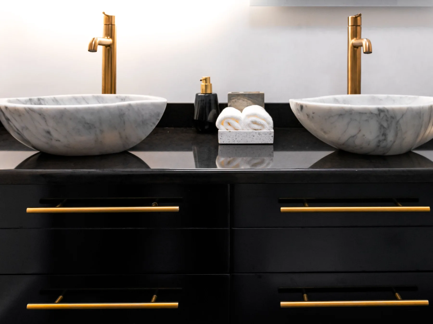 A black painted double vanity with long brass bar pulls, a black counter, two marble vessel basins and brass faucets