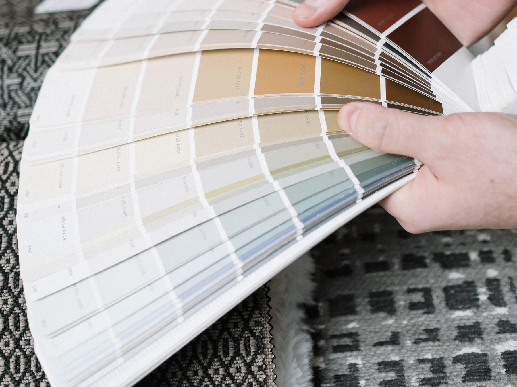 Two hands fanning out a deck of warm neutral paint colour swatches over woven fabric samples