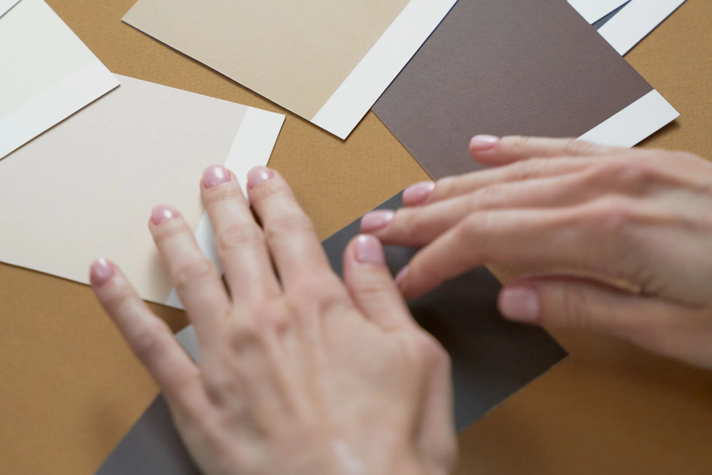 Two hands arranging neutral paint sample cards on a tan table