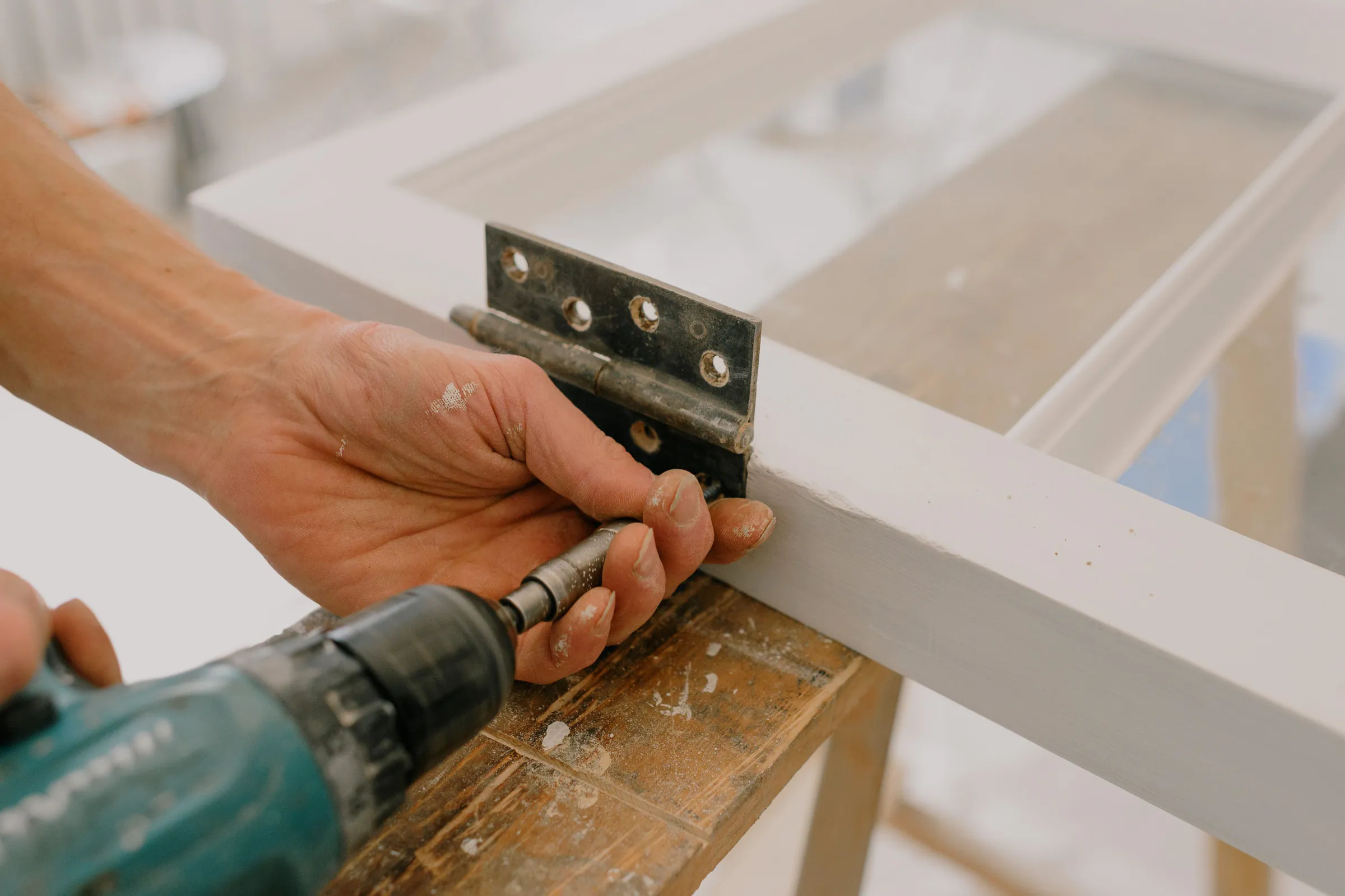 A hand holding a cordless drill driver to the screws of a metal hinge on a white painted frame