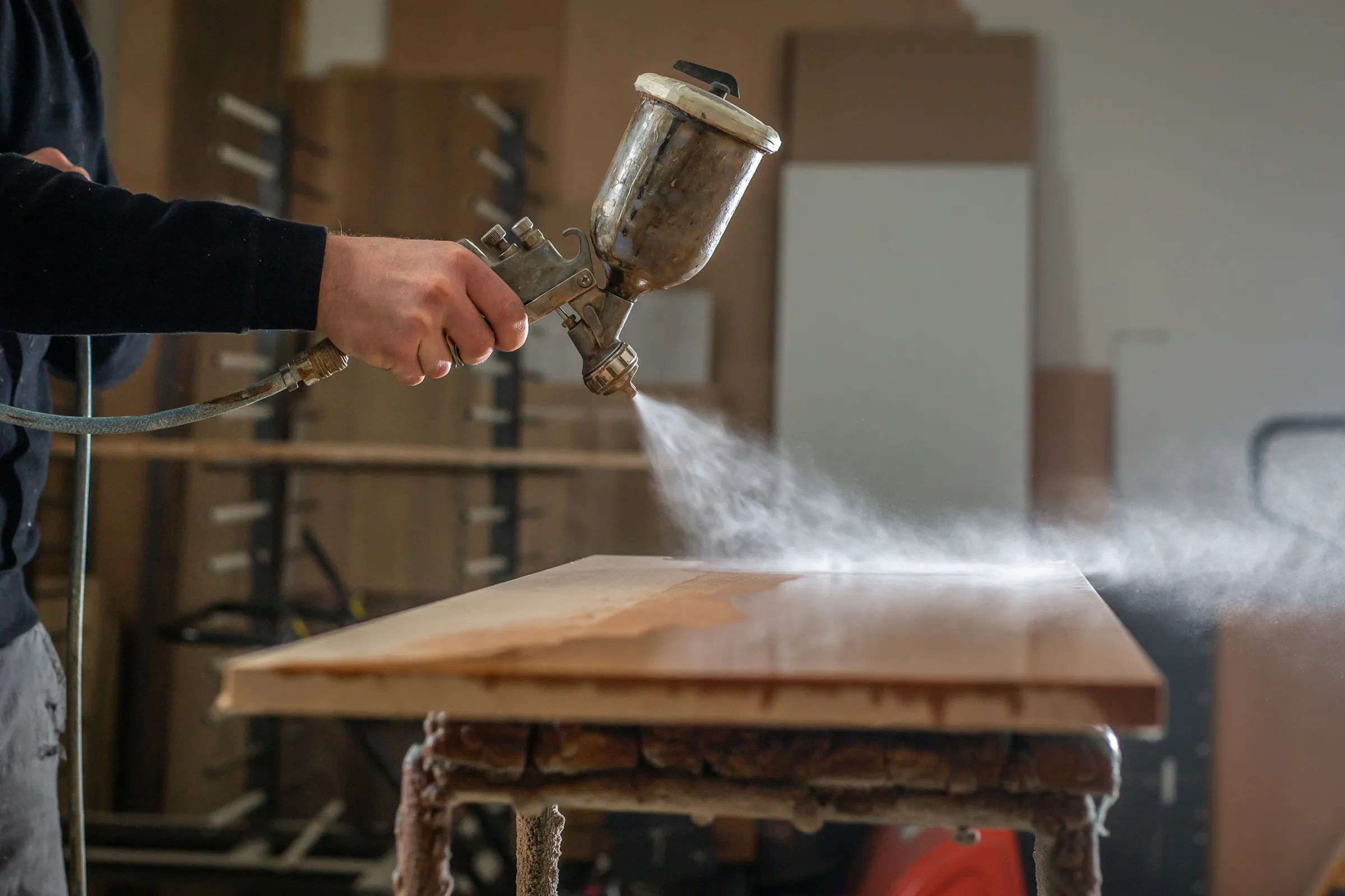 A hand-held gravity-feed spray gun misting a coat across a wooden panel in a shop, with panels stacked on racks behind