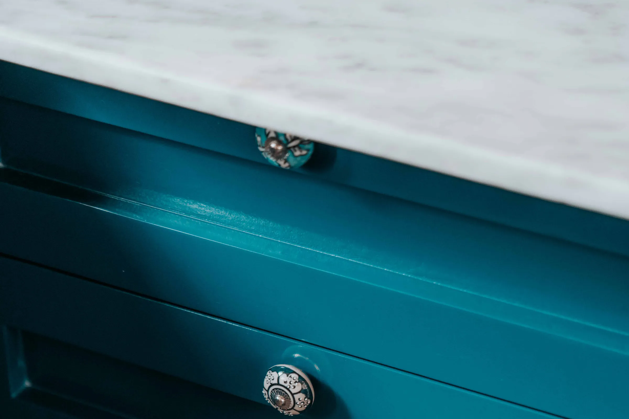 Teal painted drawer fronts with patterned round knobs under a white marble top
