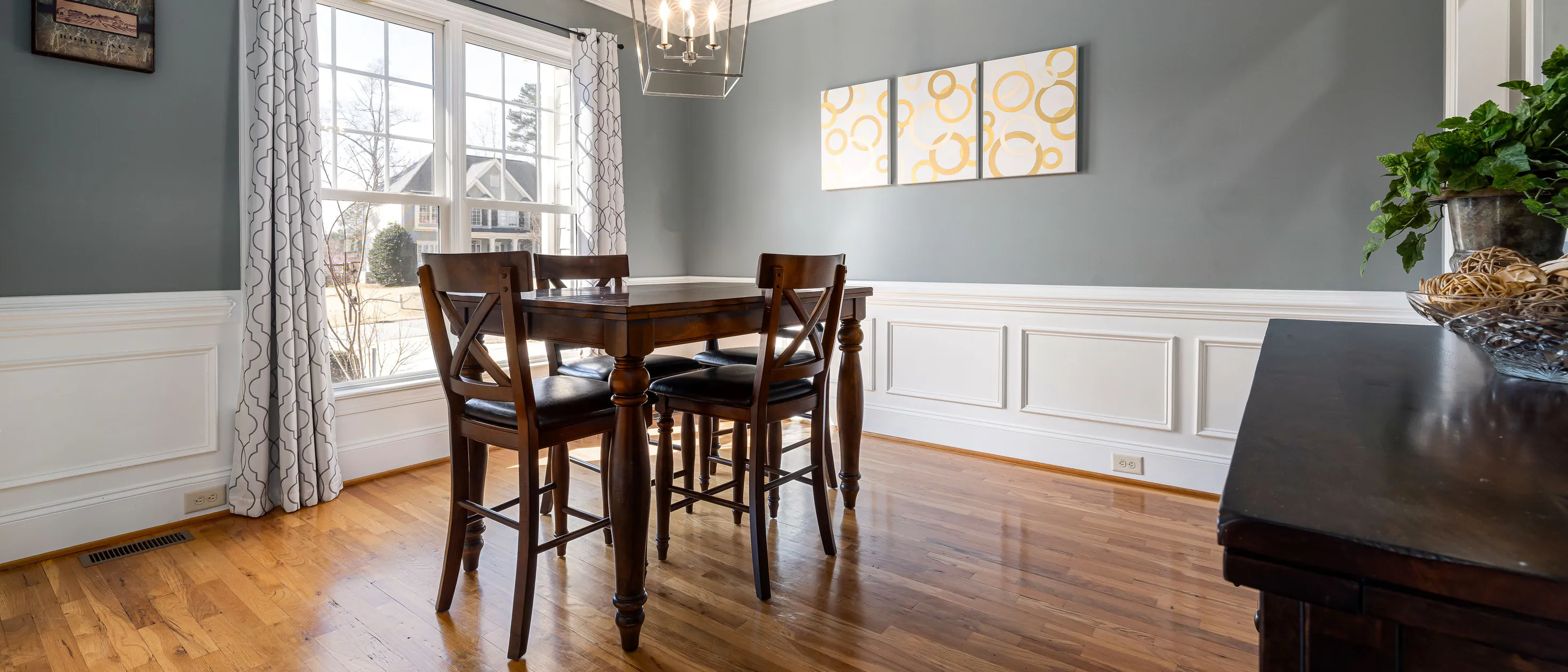 A dining room with white raised-panel wainscoting, crown moulding and grey-green walls above a wood floor