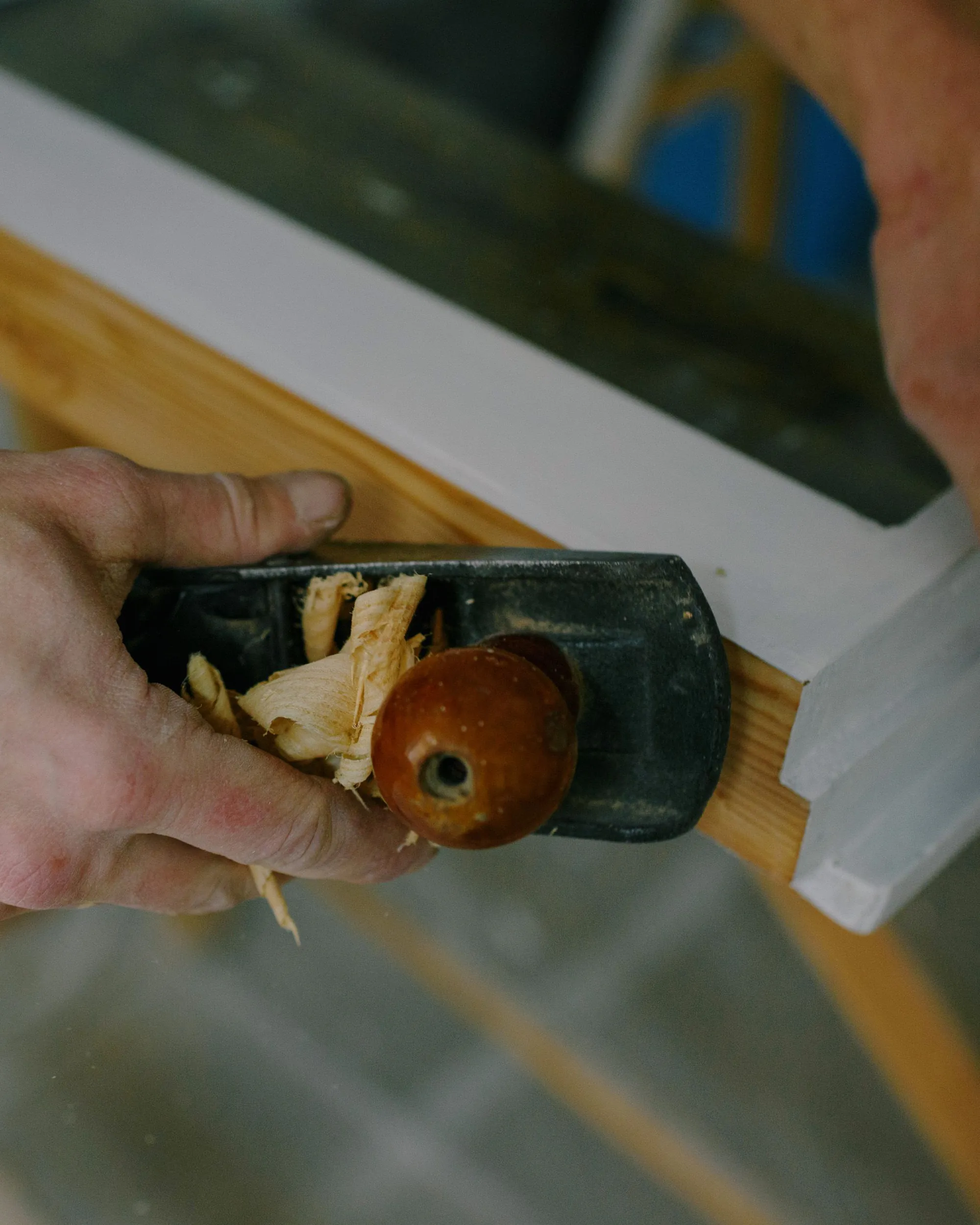 Hands running a hand plane along the edge of a white-painted wooden window frame, shavings curling off