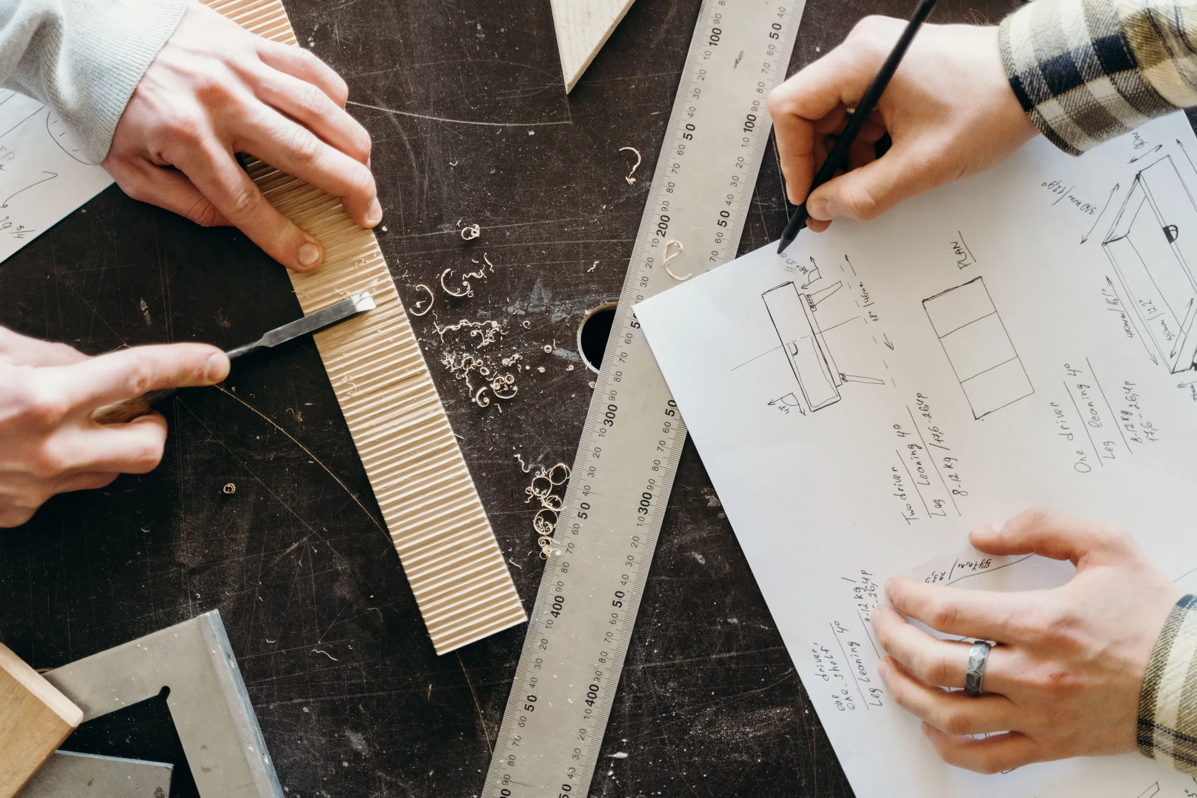 Hands sketching furniture plans on paper beside a steel ruler and a strip of wood on a workbench