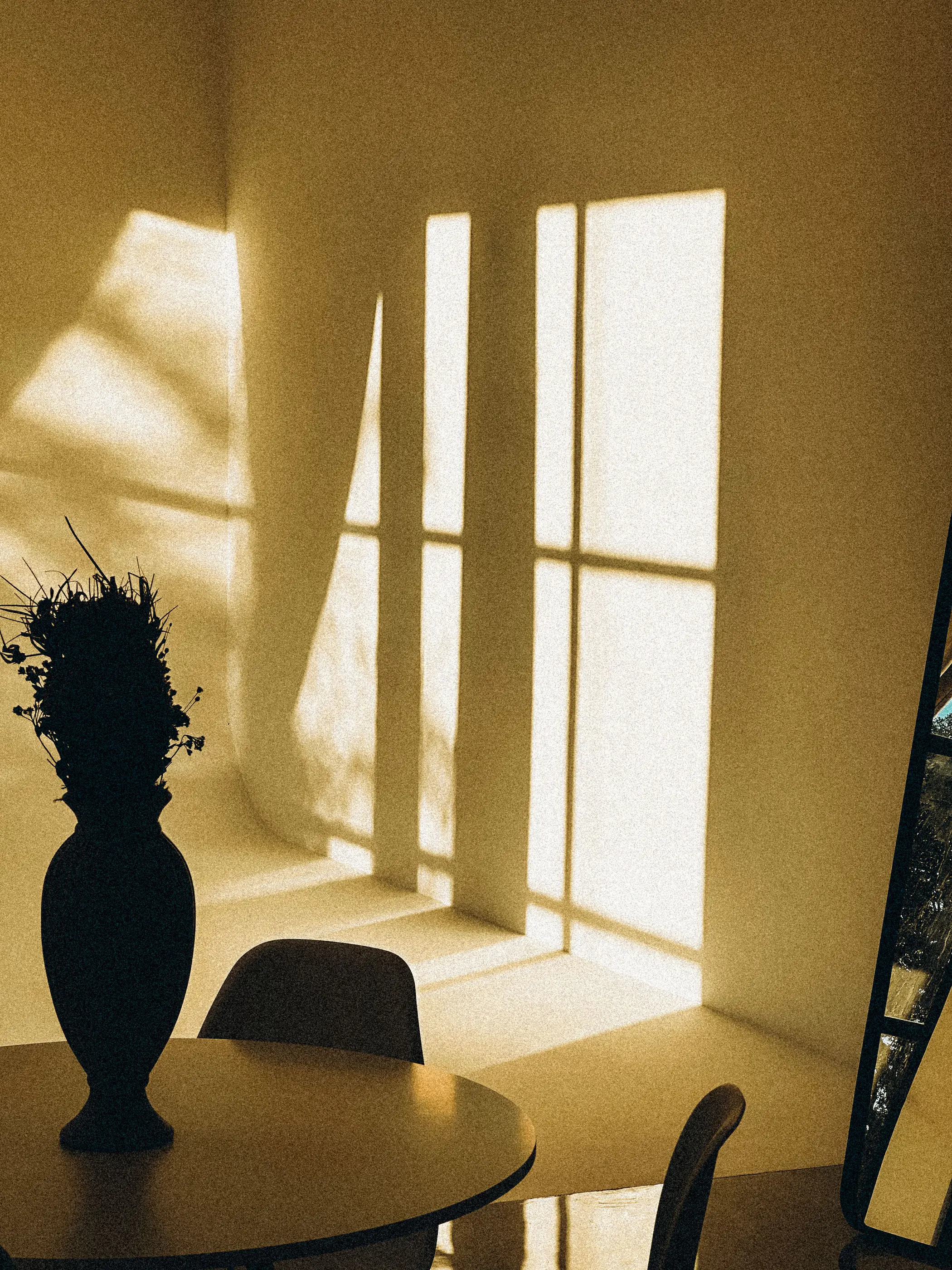 Late sun throwing window shadows across a warm yellow wall beside a dark vase on a table
