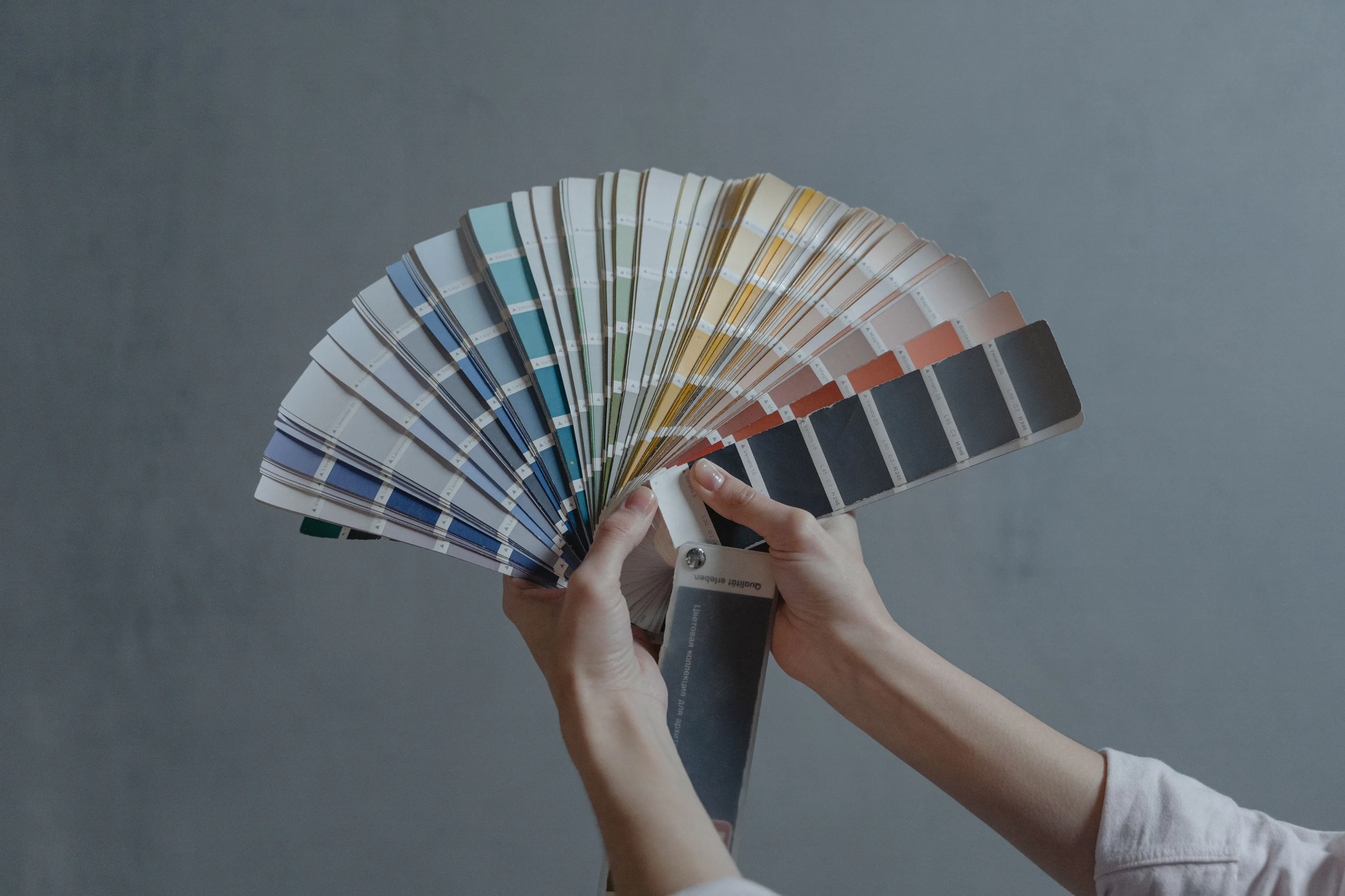 A fan deck of paint color cards held open by two hands against a gray wall