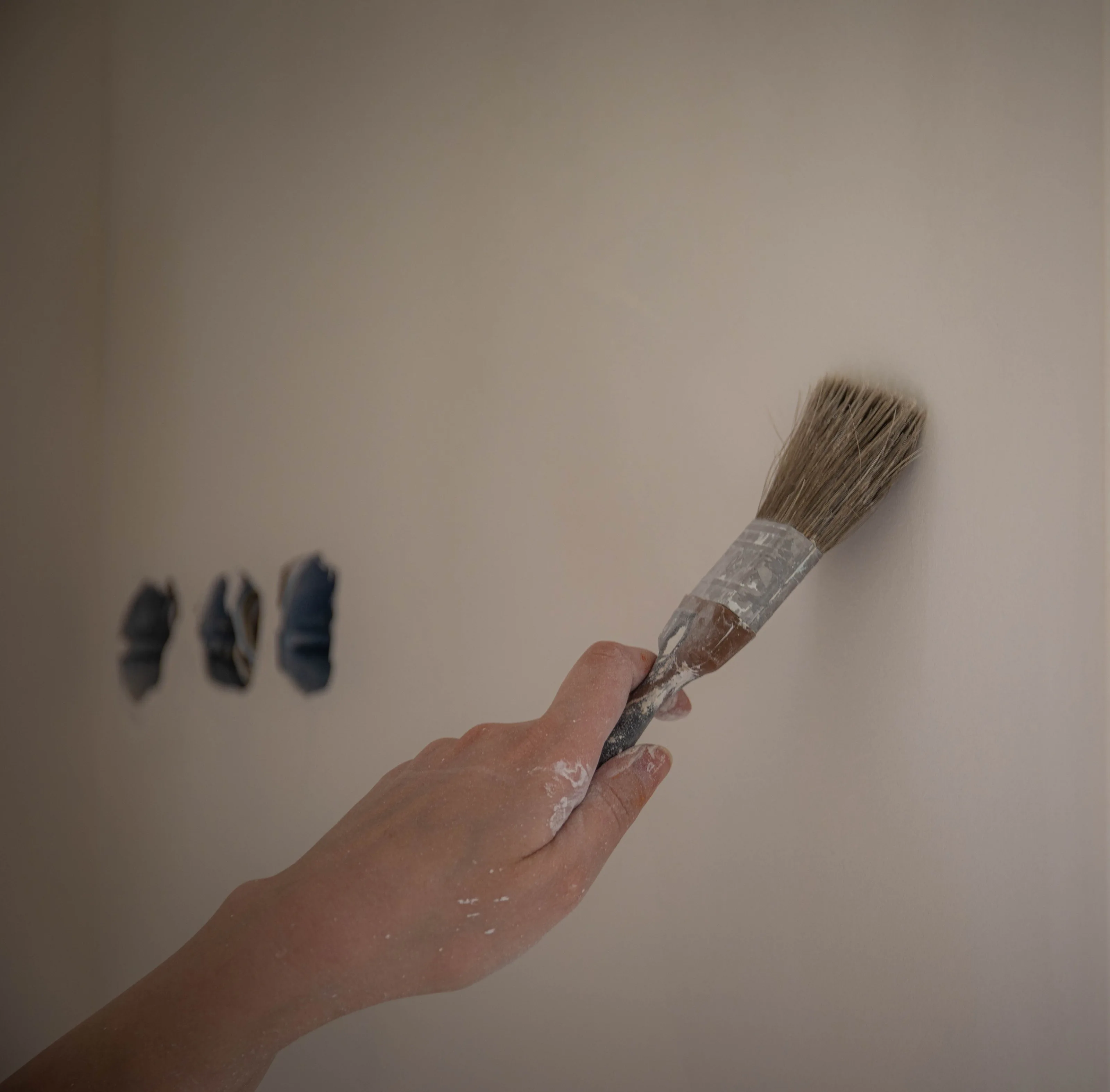 A hand brushing small trial patches of dark paint onto a pale wall