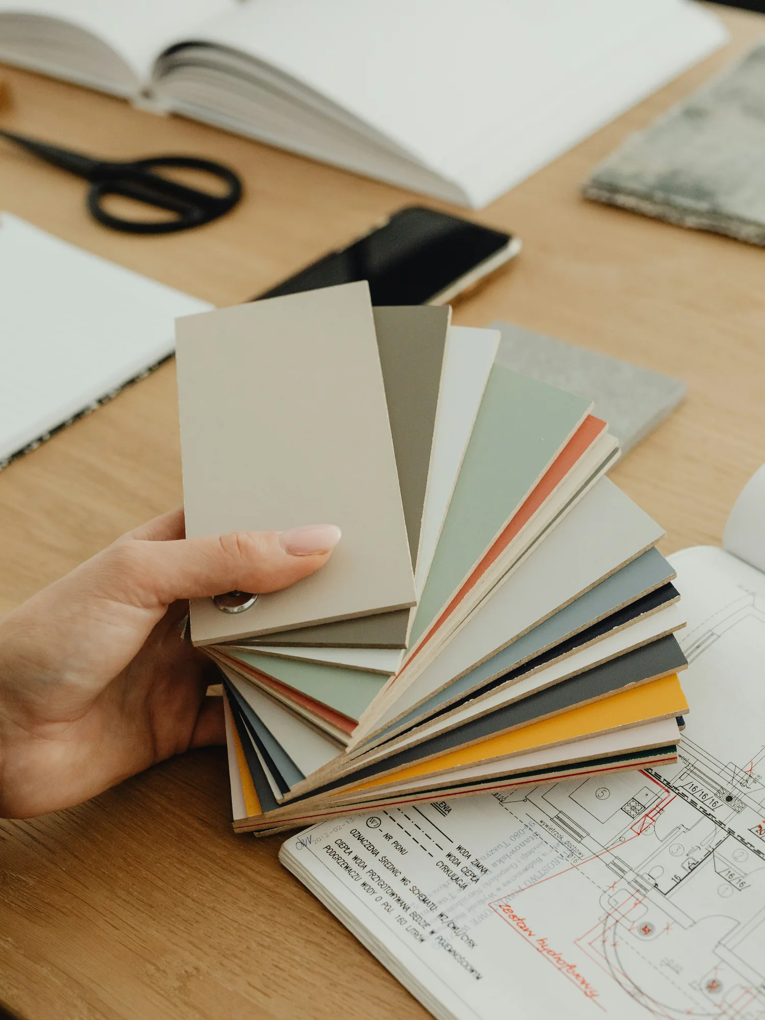 A hand fanning out painted colour sample cards over a floor plan drawing on a wooden table, beside an open notebook