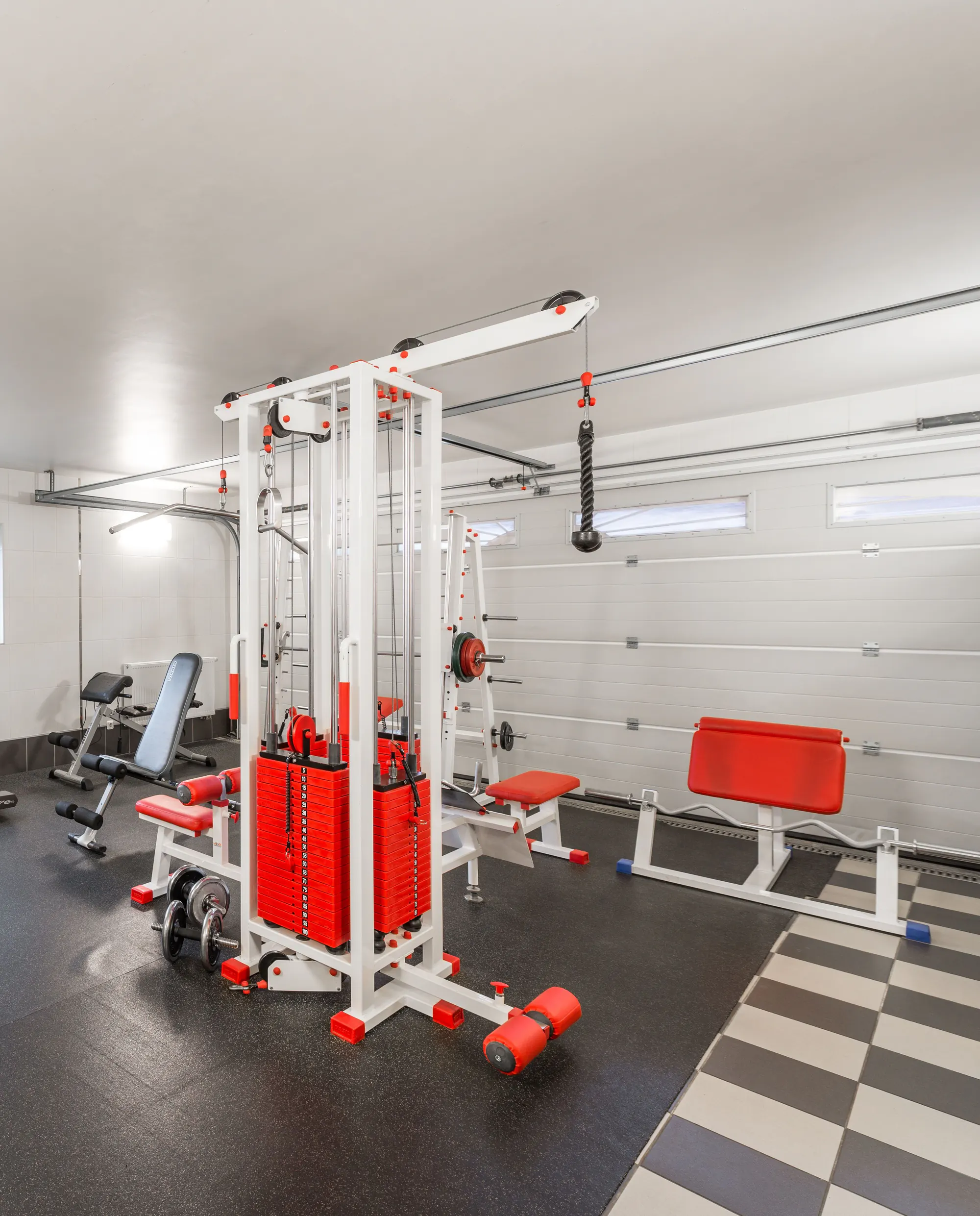 A garage fitted out as a home gym, with a white and red cable machine and benches on a black rubber floor in front of the closed garage door
