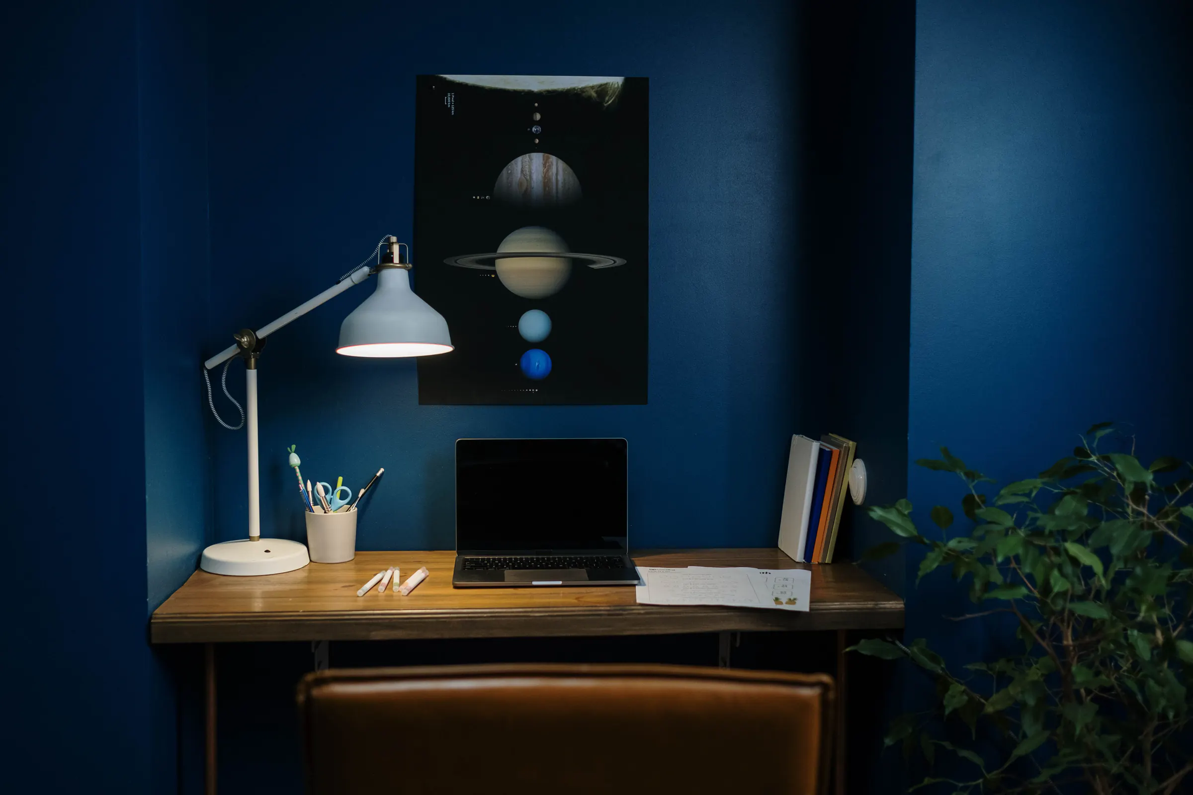 A wood desk with a laptop and a white lamp against a deep navy wall, under a poster of the planets